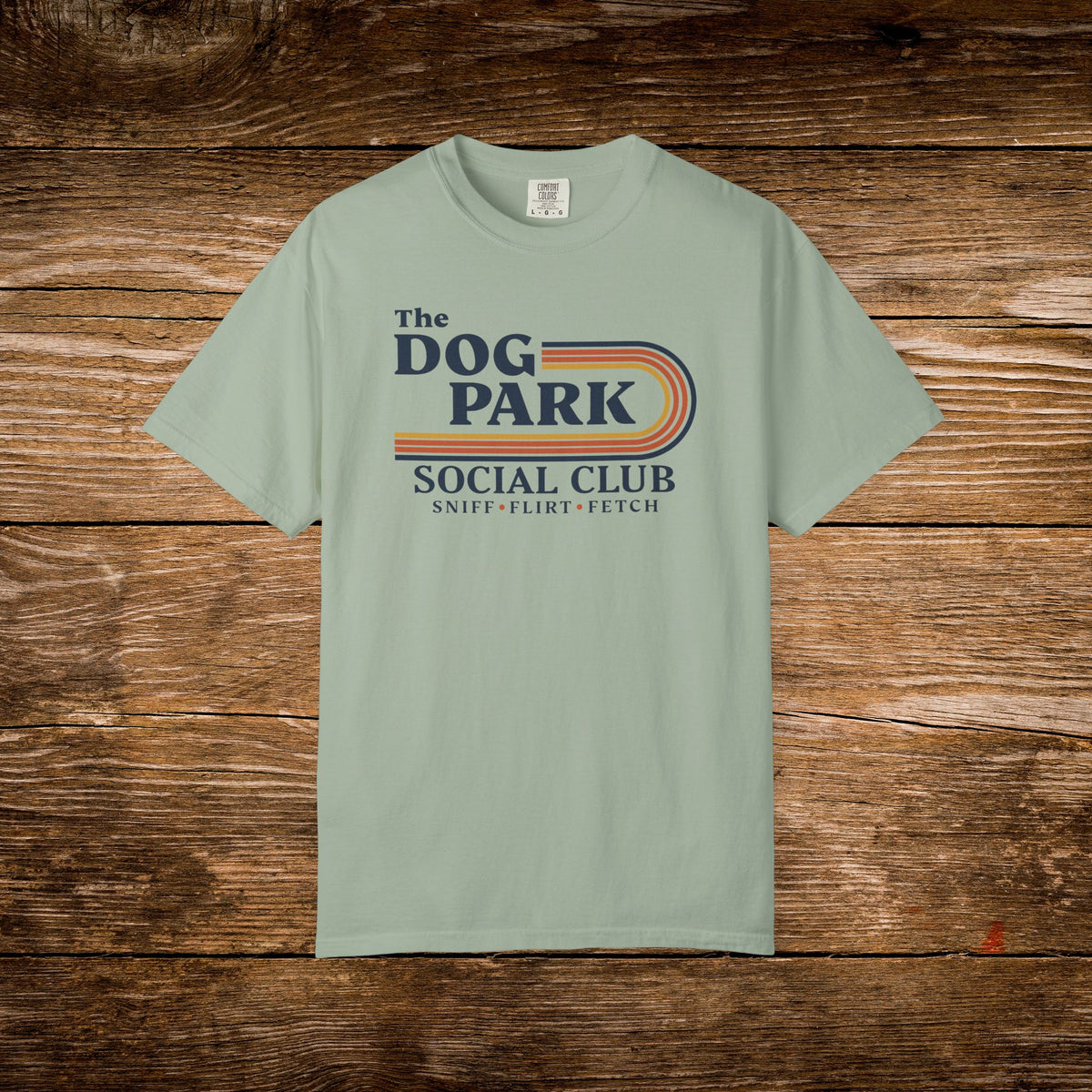 A sage green short-sleeve t-shirt with a retro-style graphic reading 'The DOG PARK SOCIAL CLUB' and 'SNIFF - FLIRT - FETCH' in navy and orange, laid flat on a rustic brown wooden background.