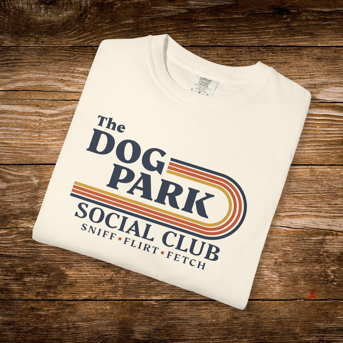 A folded off-white t-shirt with a retro-style graphic reading 'The DOG PARK SOCIAL CLUB' and 'SNIFF • FLIRT • FETCH' in navy and orange-yellow stripes, placed on a rustic wooden surface.