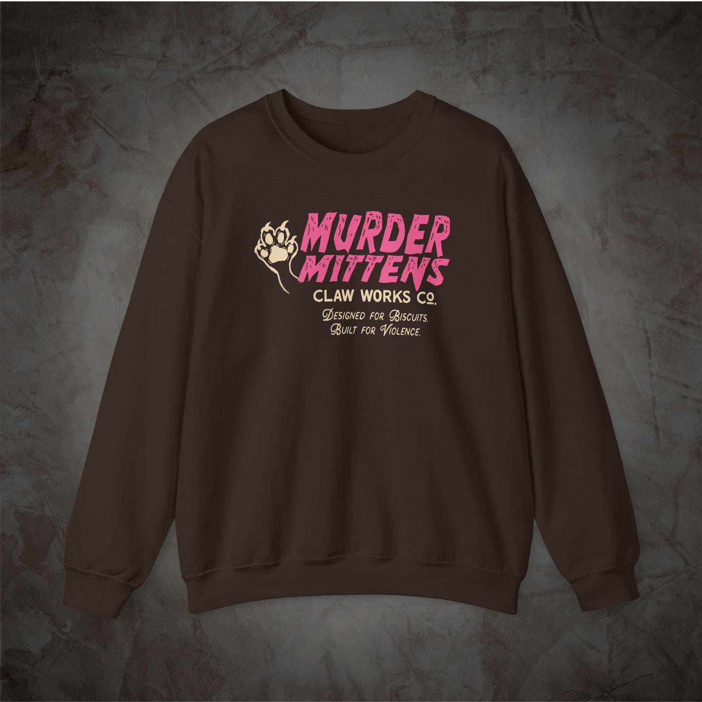 A dark brown crewneck sweatshirt featuring a graphic print on the chest with the words 'MURDER MITTENS CLAN WORKS CO.' in pink and beige, displayed on a textured gray background.
