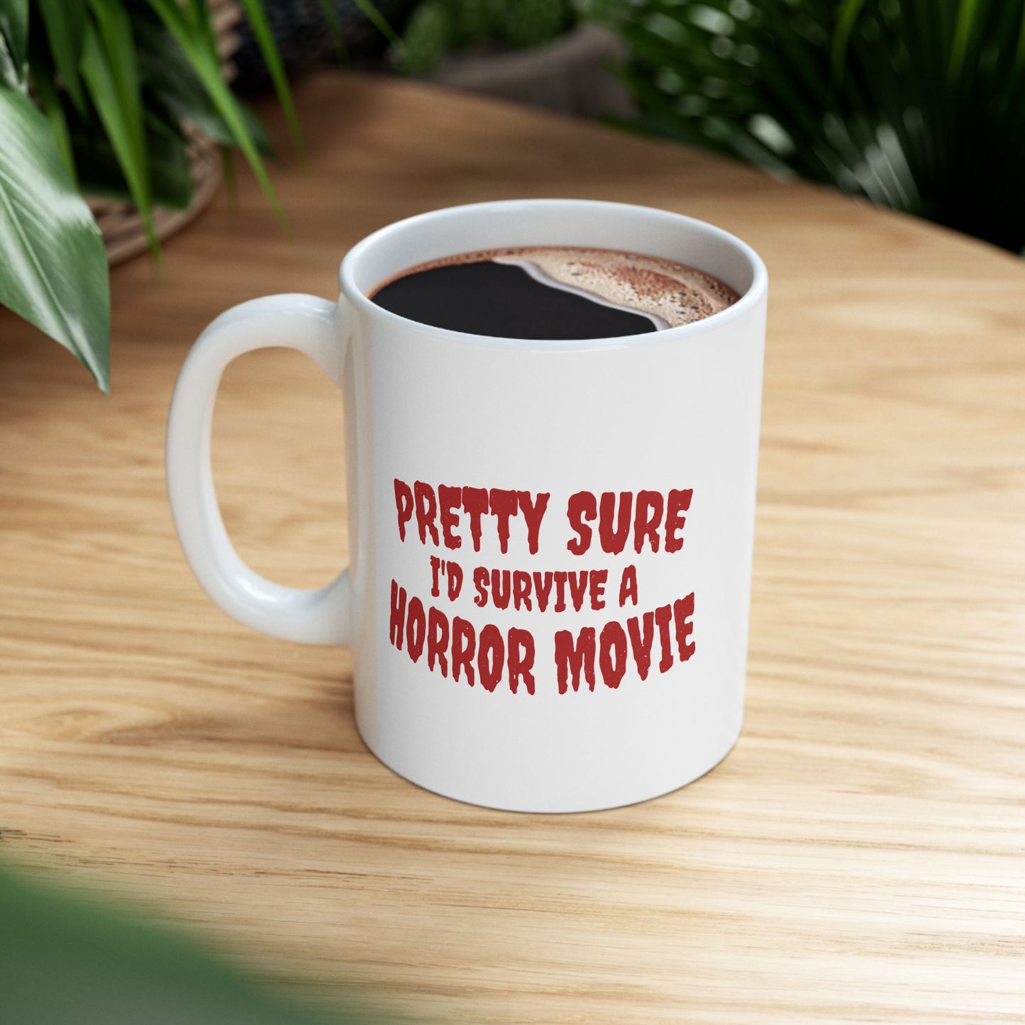 A white ceramic coffee mug with red text reading 'PRETTY SURE I'D SURVIVE A HORROR MOVIE' sits on a light wood table with green plants blurred in the background.