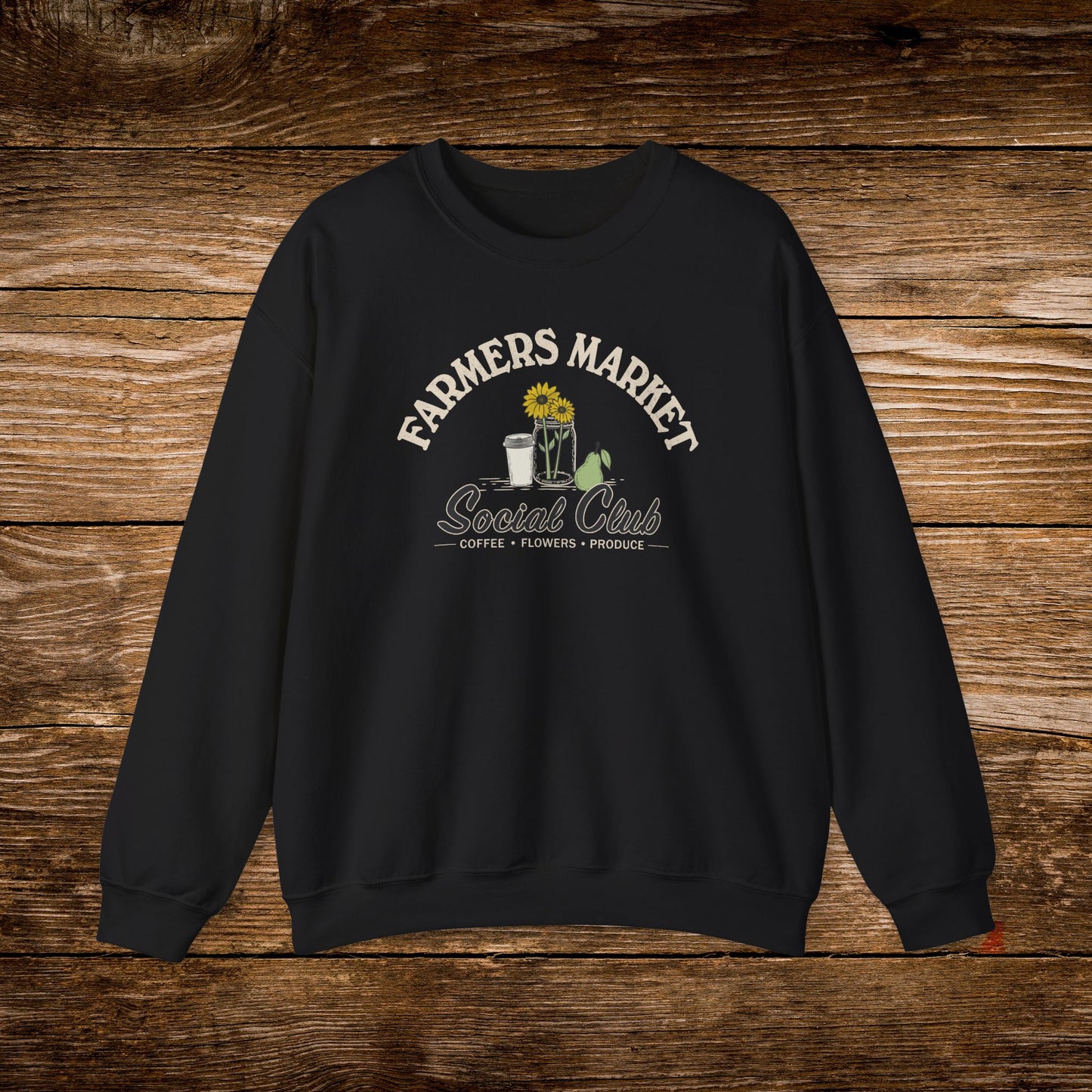 A black crewneck sweatshirt with a vintage-style graphic reading 'FARMERS MARKET Social Club' and 'COFFEE • FRUITS • PRODUCE' beneath it, featuring a sunflower and coffee cup illustration. The sweatshirt is displayed flat against a rustic brown wooden plank background.