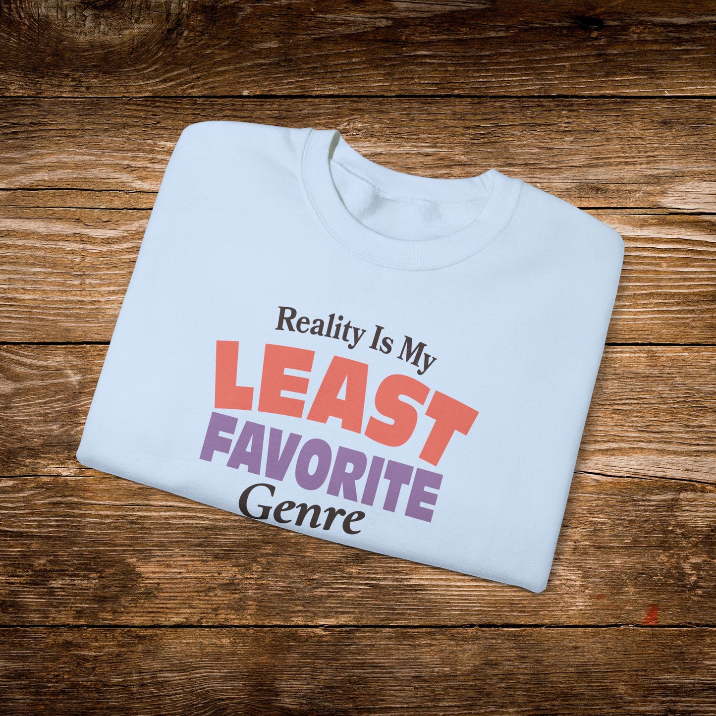 A light blue folded t-shirt with the text 'Reality Is My LEAST FAVORITE Genre' printed on the front, displayed on a rustic brown wooden background.