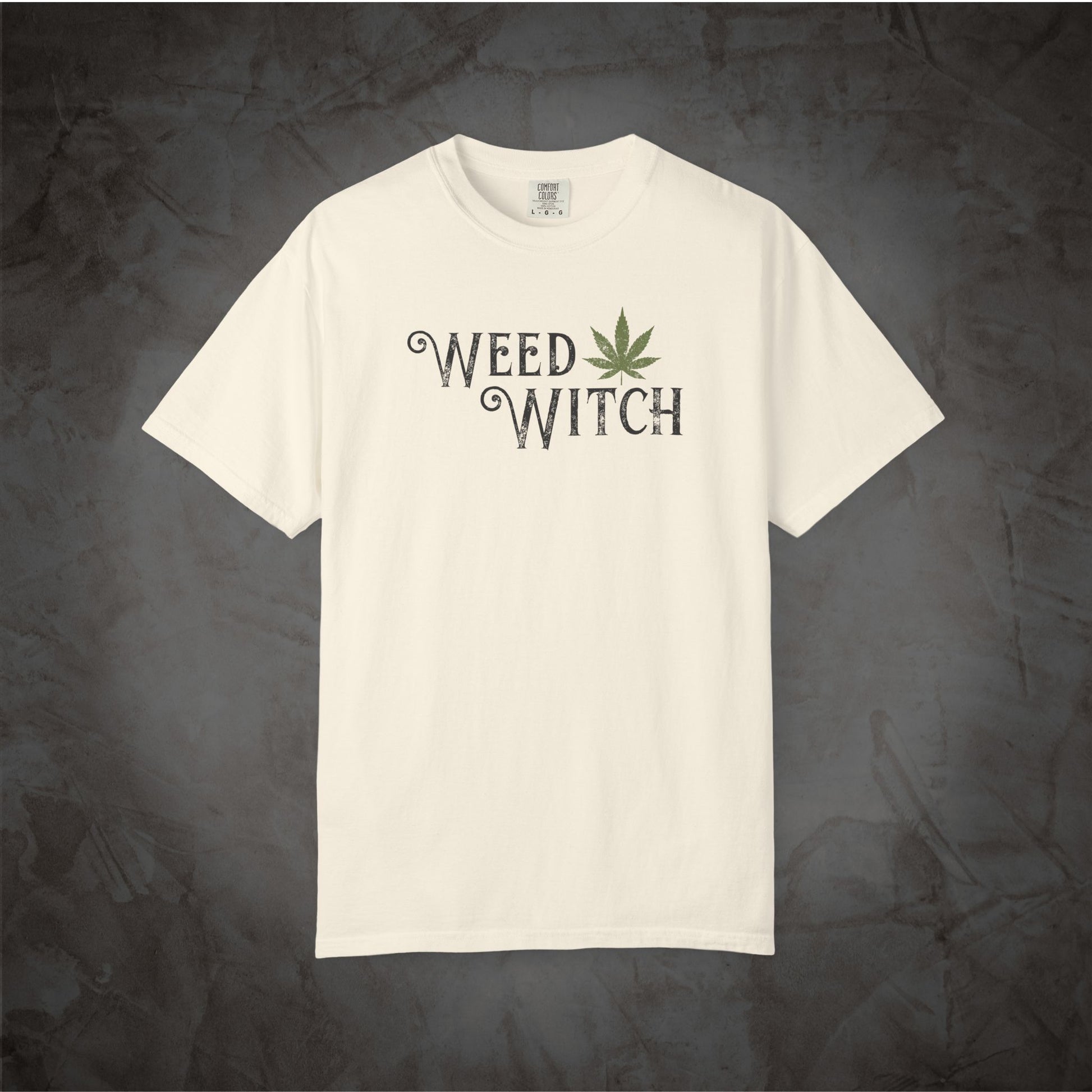 An off-white short-sleeve t-shirt with a distressed graphic on the chest reading 'WEED WITCH' in black, featuring a green cannabis leaf as the letter 'I'. The t-shirt is displayed flat against a dark, textured gray background.