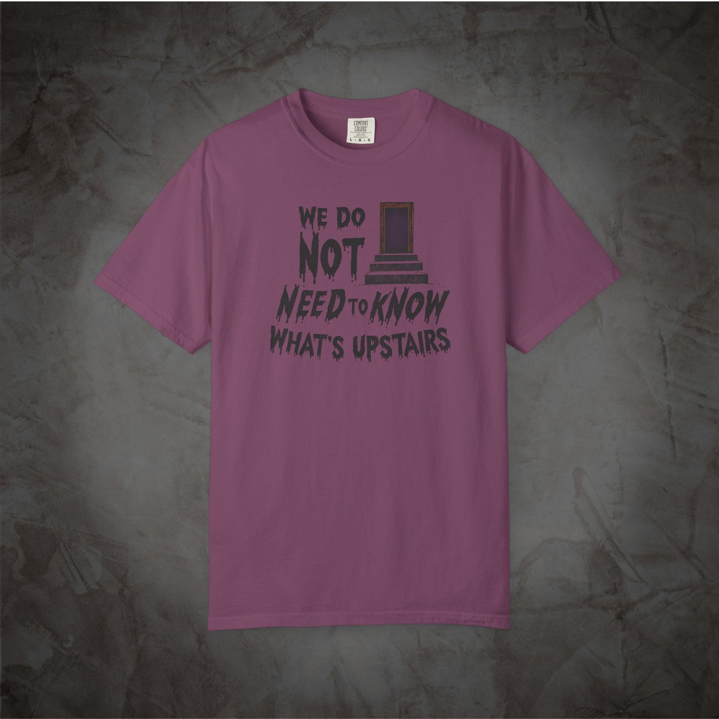 A mauve-purple short-sleeve t-shirt with a dark graphic print on the front reading 'WE DO NOT NEED TO KNOW WHAT'S UPSTAIRS' alongside a depiction of the TARDIS door, displayed flat against a textured gray background.