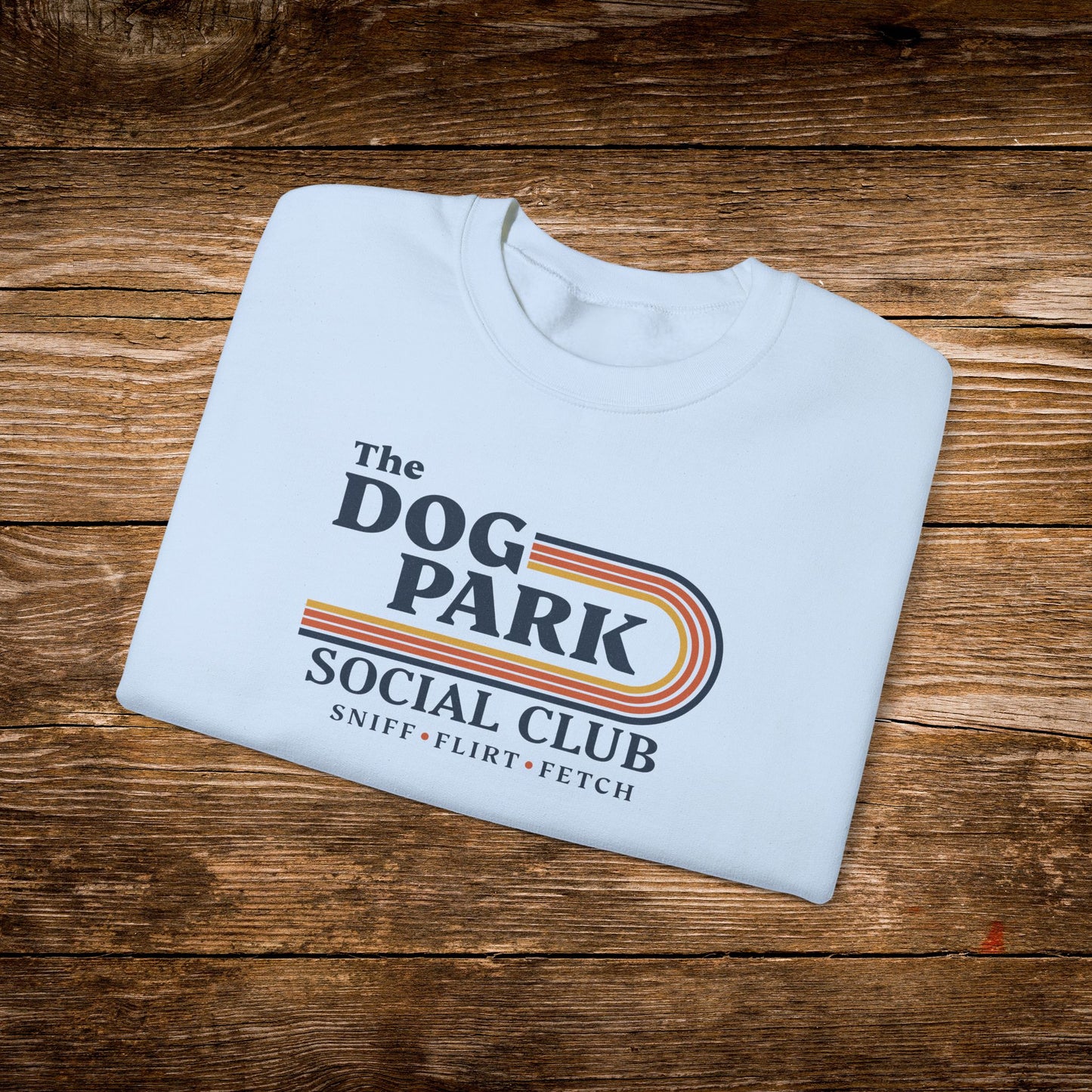 A folded white t-shirt with a colorful retro-style graphic reading 'The Dog Park Social Club' and 'Sniff, Flirt, Fetch' lies on a rustic brown wooden plank background.