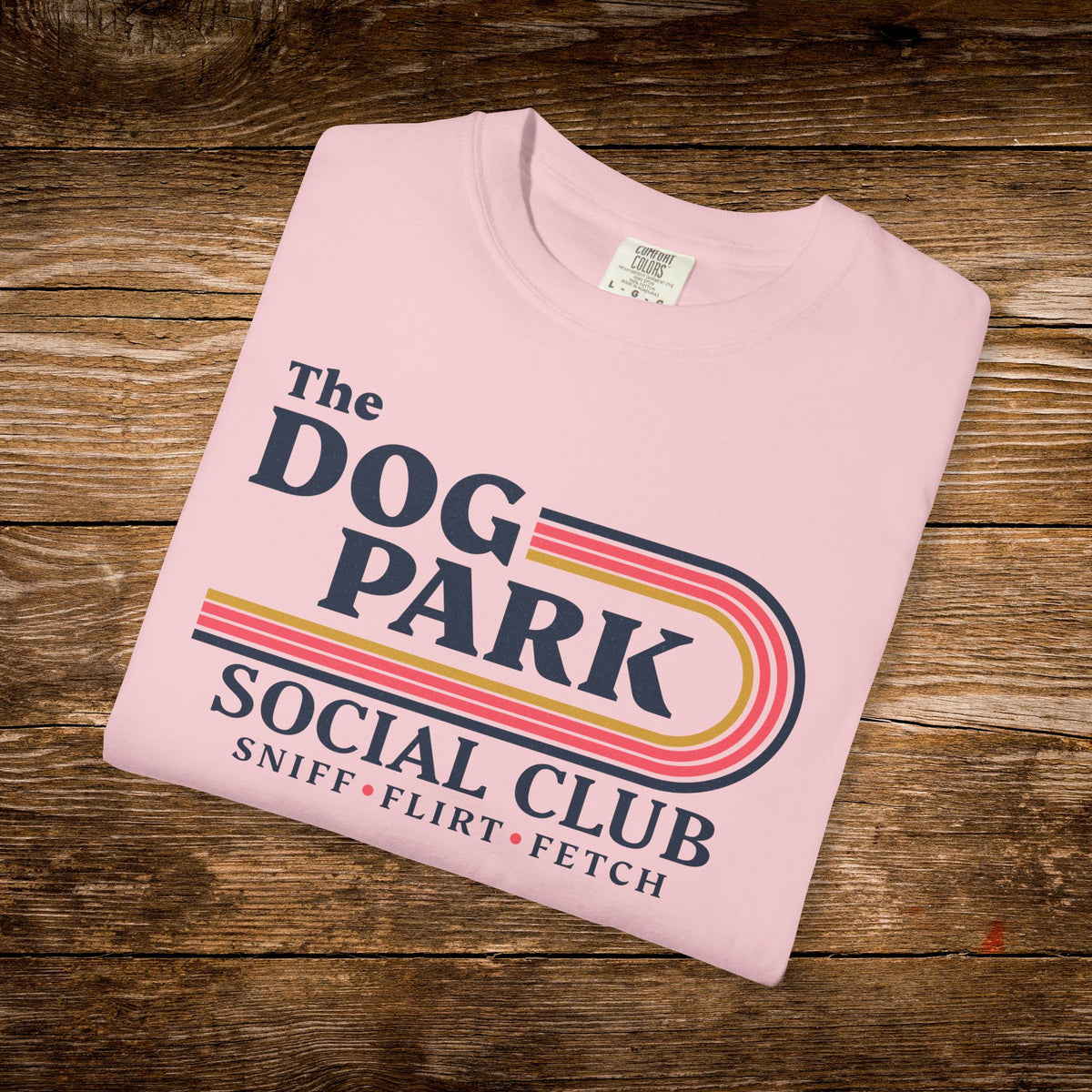 A folded light pink t-shirt with a retro-style 'The Dog Park Social Club' graphic, displayed on a rustic wooden background.