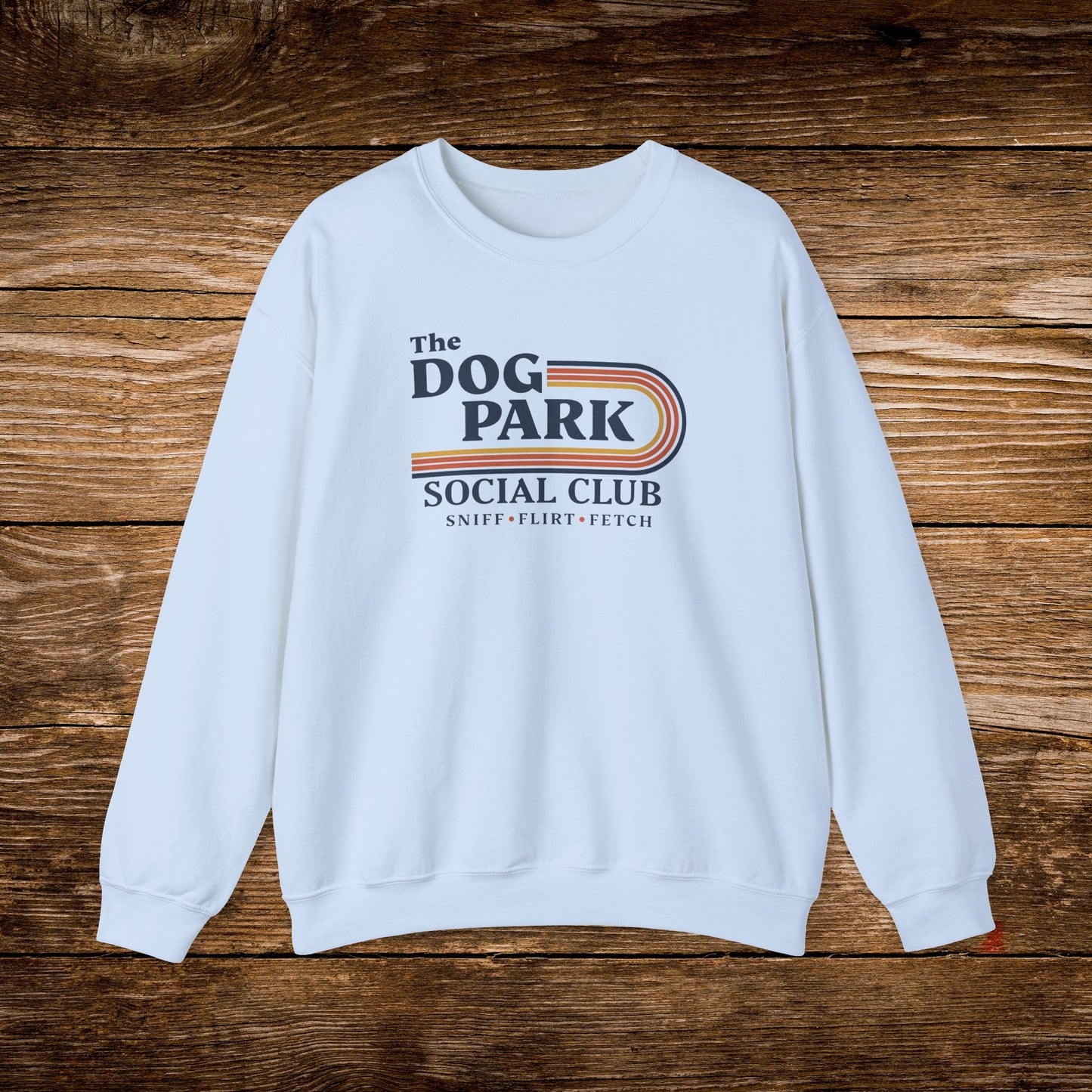 A light blue crewneck sweatshirt with a retro-style graphic reading 'The Dog Park Social Club' and 'Sniff • Flirt • Fetch' in navy, orange, and beige, laid flat on a rustic brown wooden background. The design text reads "Sniff, Flirt, Fetch."