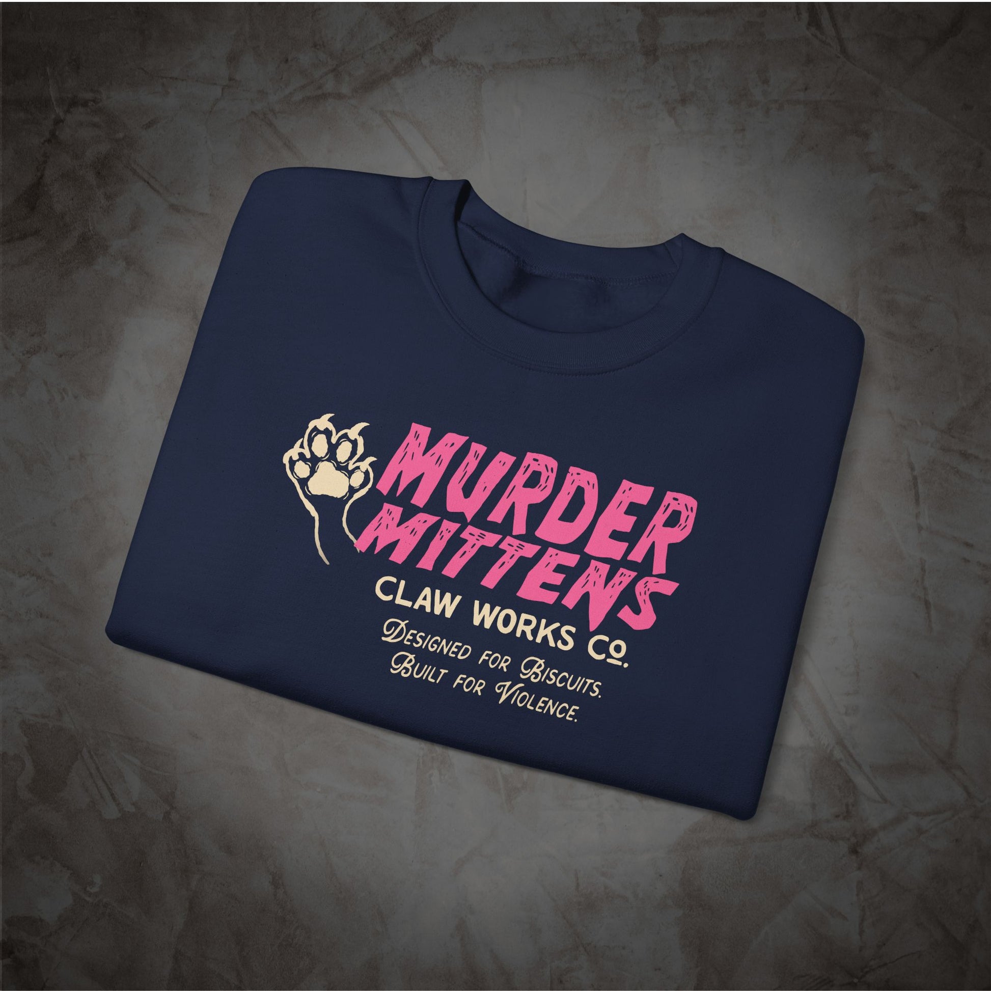 A navy blue folded t-shirt with a pink and white graphic print reading 'MURDER MITTENS CLAW WORKS CO. DESIGNED FOR BISCUITS.