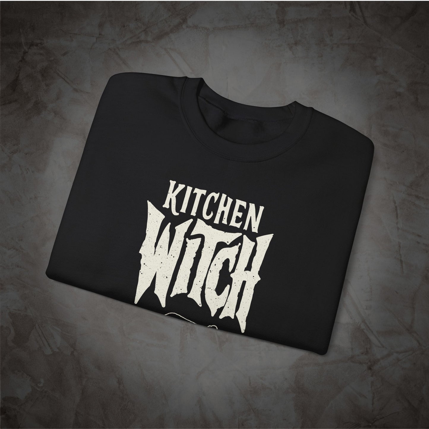 A black crewneck t-shirt with a distressed white graphic reading 'KITCHEN WITCH' in a stylized gothic font, displayed flat on a textured gray background.