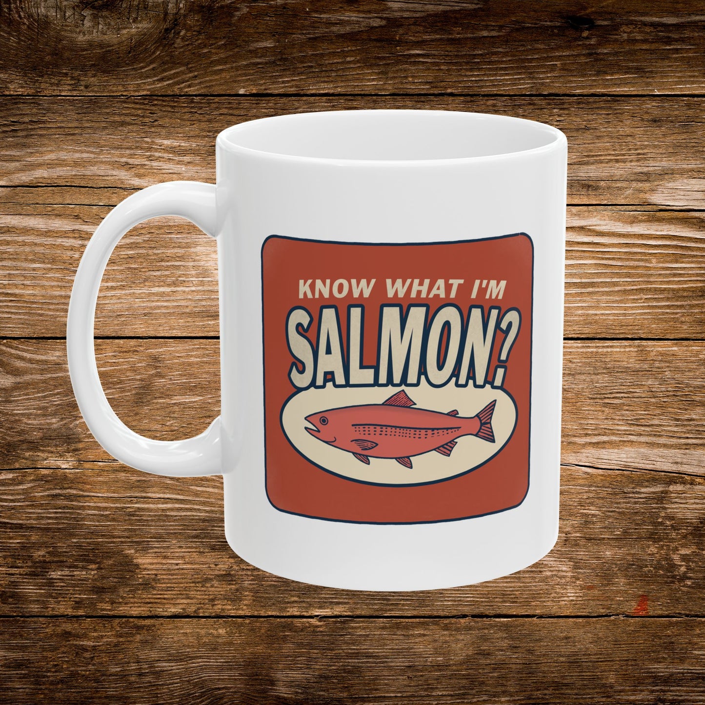 A white ceramic coffee mug with a red and beige graphic that reads 'KNOW WHAT I'M SALMON?' and shows an illustration of a salmon, set against a rustic wooden background.