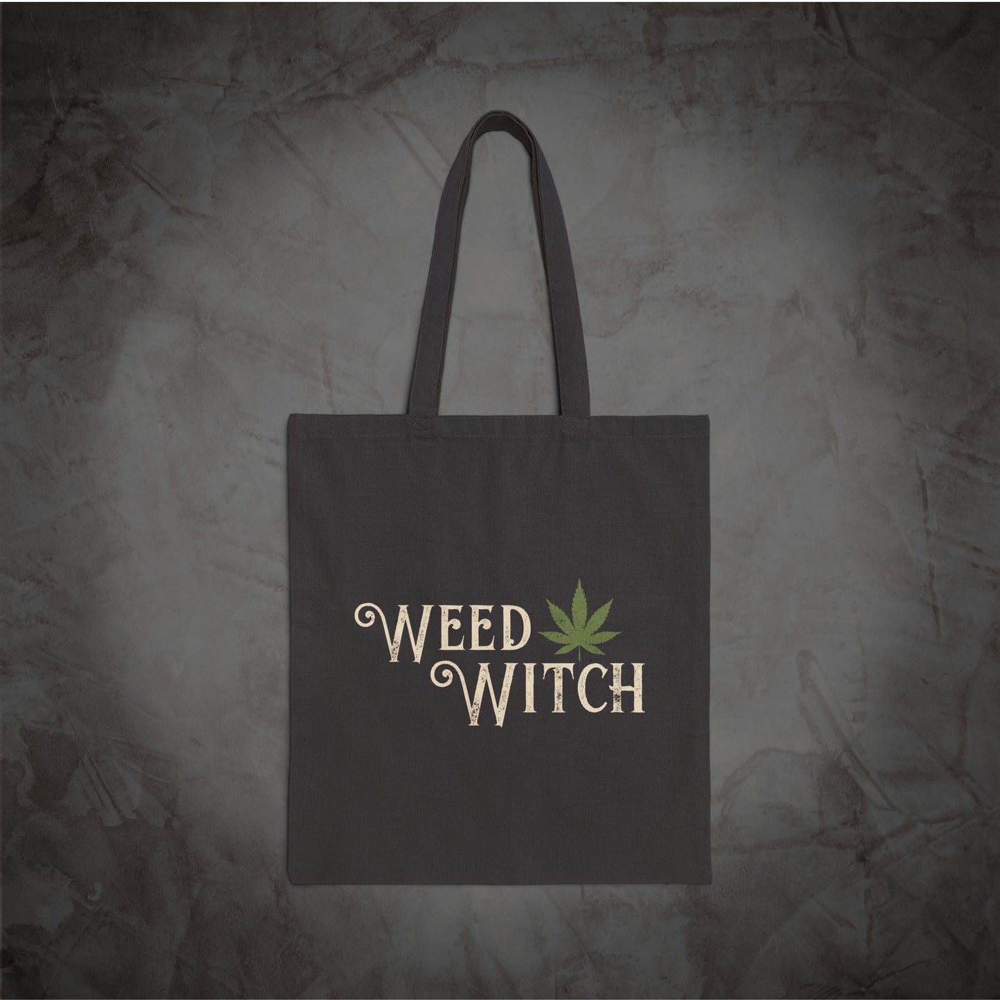 A dark gray canvas tote bag with two handles, featuring the words 'WEED WITCH' in a beige gothic font and a green cannabis leaf graphic, set against a textured gray background.