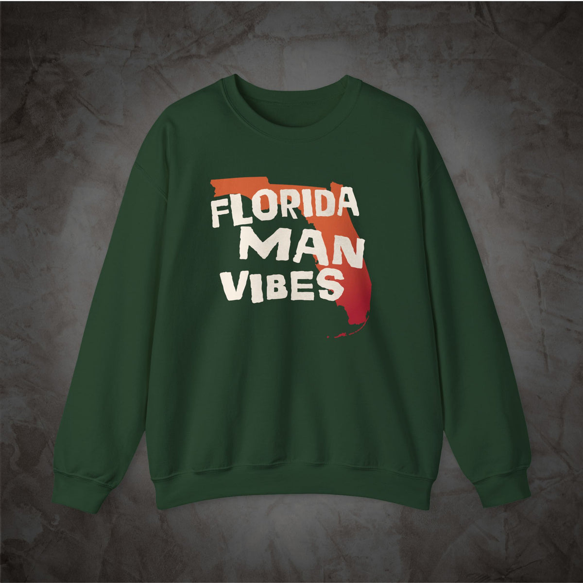 A dark green crewneck sweatshirt featuring a graphic on the chest with the words 'FLORIDA MAN VIBES' in white over a red and orange Florida state outline, displayed against a textured gray background.