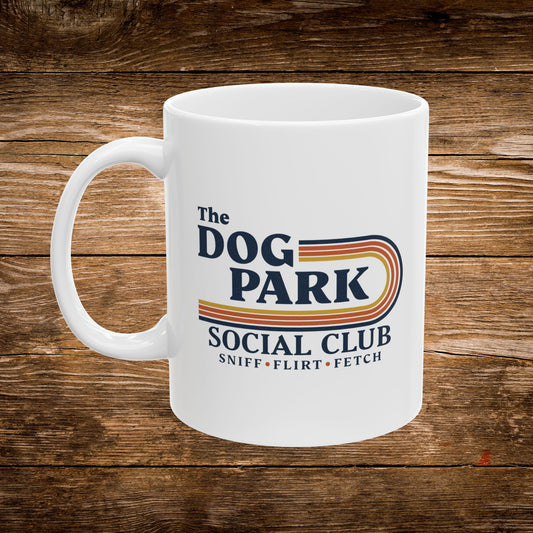 A white ceramic coffee mug with a colorful retro-style graphic that reads 'The DOG PARK SOCIAL CLUB' and 'SNIFF • FLIRT • FETCH' on a rustic brown wooden background.