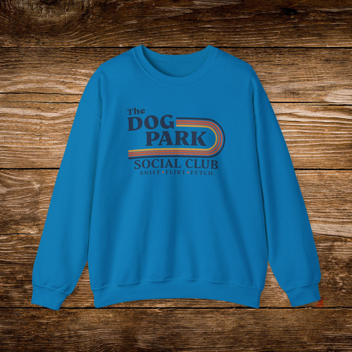 A bright blue crewneck sweatshirt with a retro-style graphic reading 'The Dog Park Social Club' and 'Sweet Flatbread T-Shirt' in dark blue and orange, displayed flat on a rustic brown wooden background.