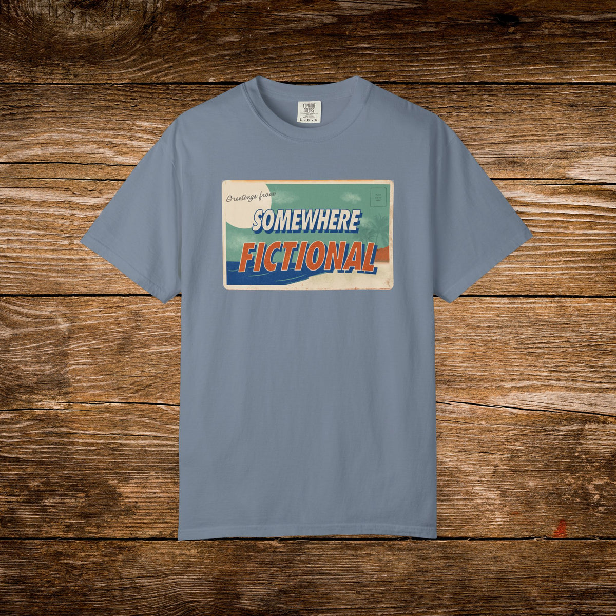 Blue Jean Comfort Colors t-shirt featuring a vintage postcard-style “Greetings from Somewhere Fictional” graphic.