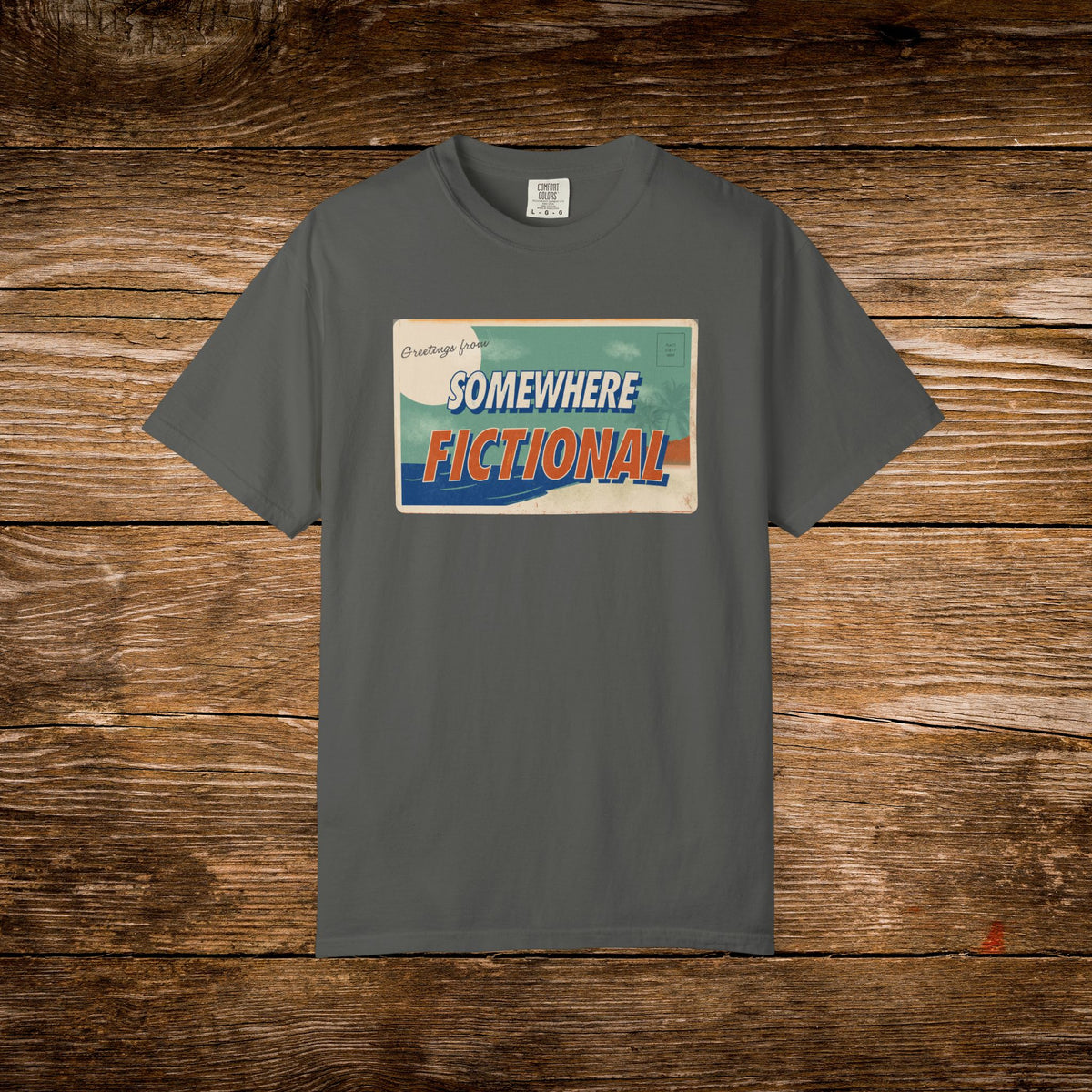Pepper Comfort Colors t-shirt with a retro “Greetings from Somewhere Fictional” postcard design.