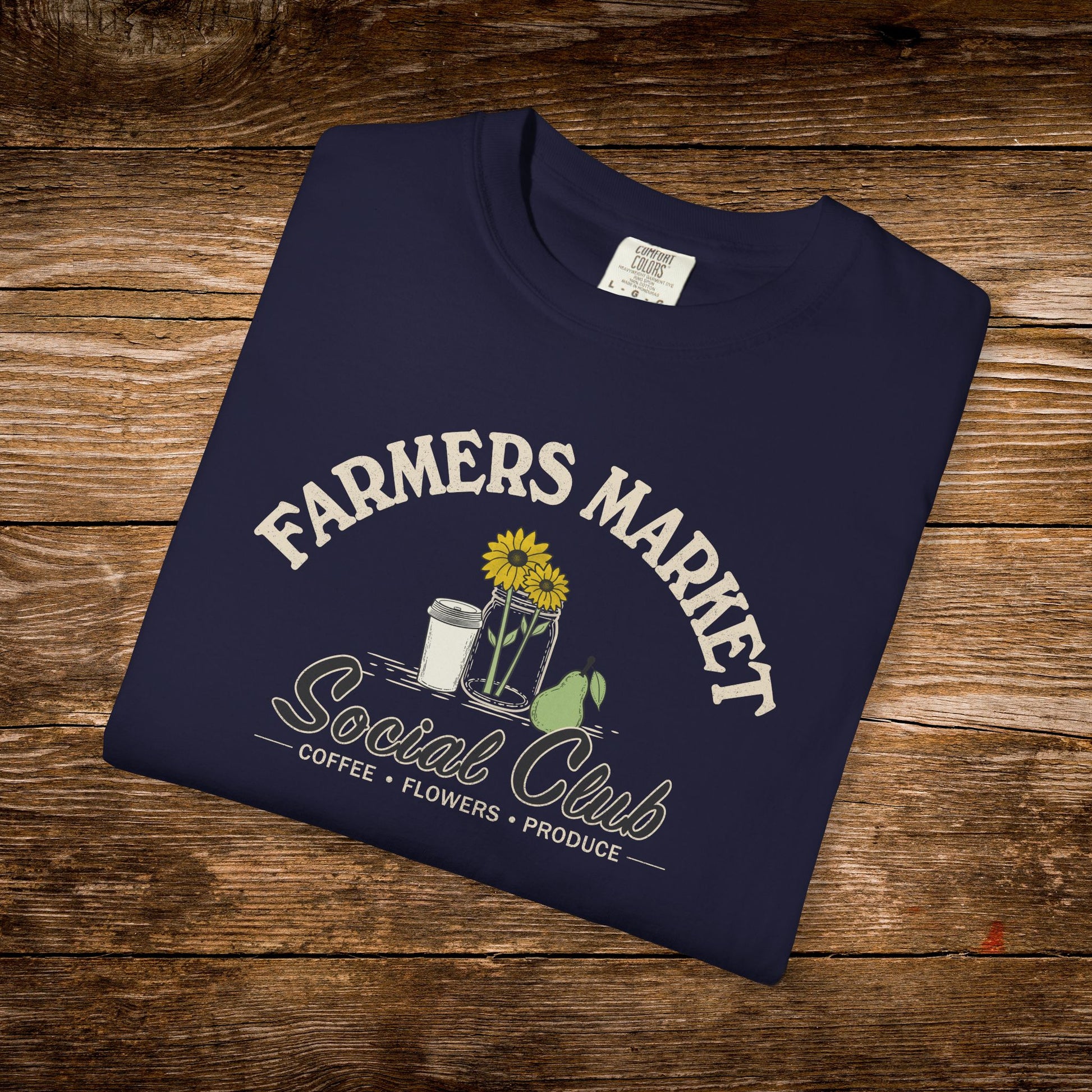 A navy blue folded t-shirt with a 'Farmers Market Social Club' graphic, displayed on a rustic wooden background.