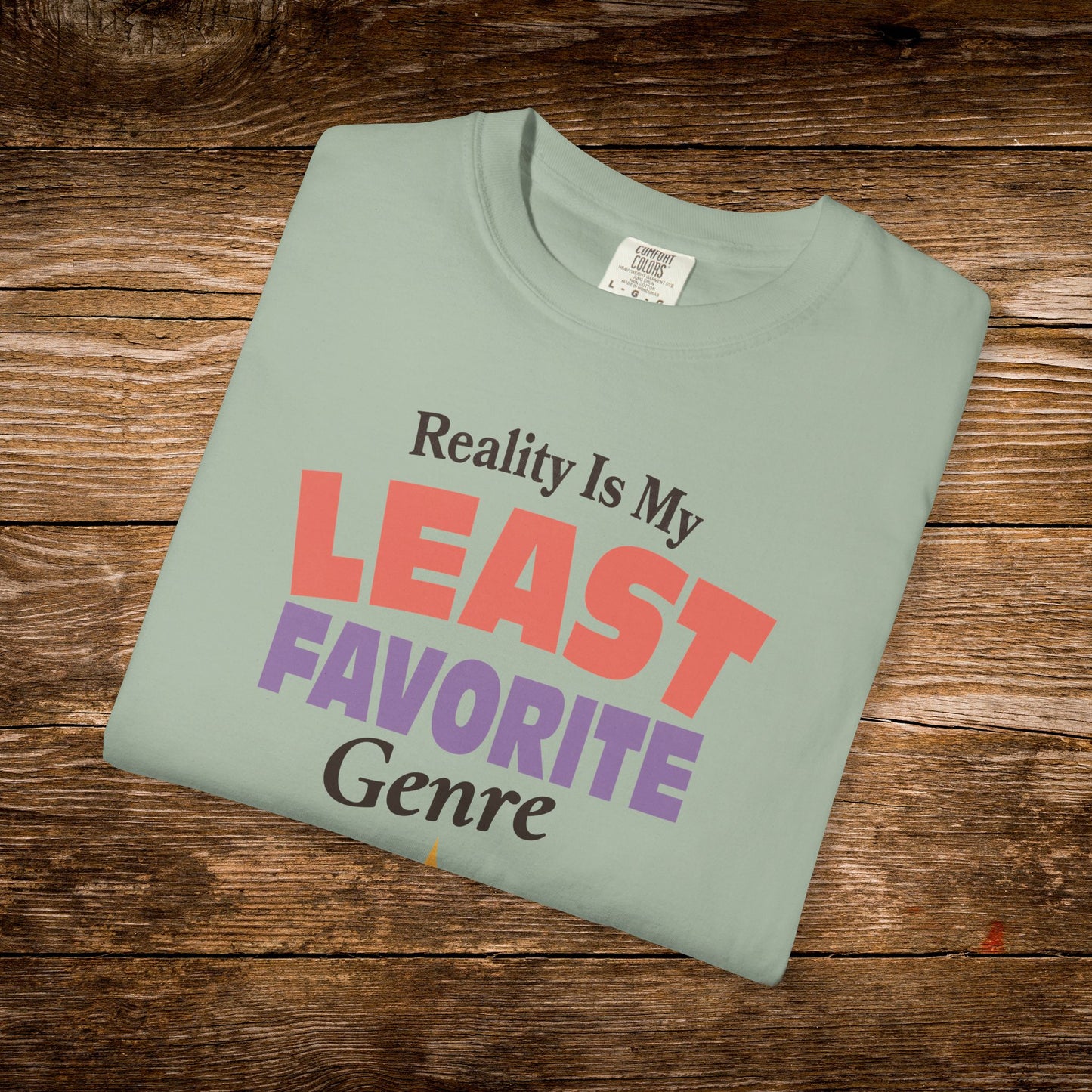 A folded sage green t-shirt with the text 'Reality Is My LEAST FAVORITE Genre' printed on the front, displayed on a rustic wooden background.