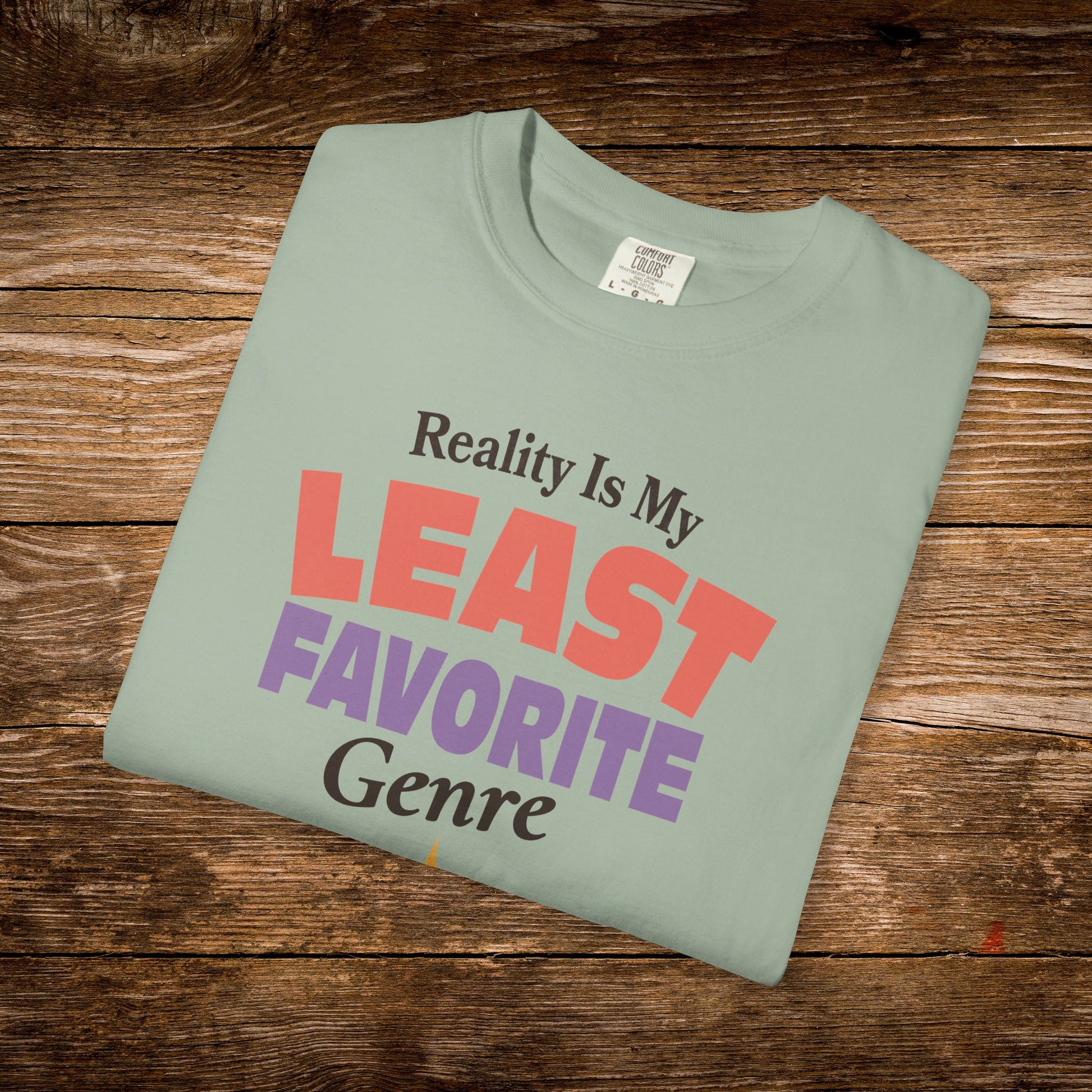 A folded sage green t-shirt with the text 'Reality Is My LEAST FAVORITE Genre' printed on the front, displayed on a rustic wooden background.
