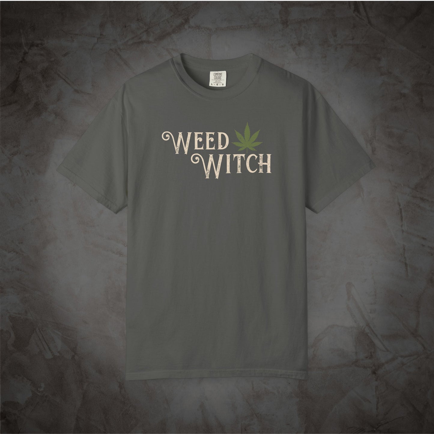 A dark gray crewneck t-shirt featuring the words 'WEED WITCH' in a stylized beige font with a green cannabis leaf graphic, displayed against a textured gray background.