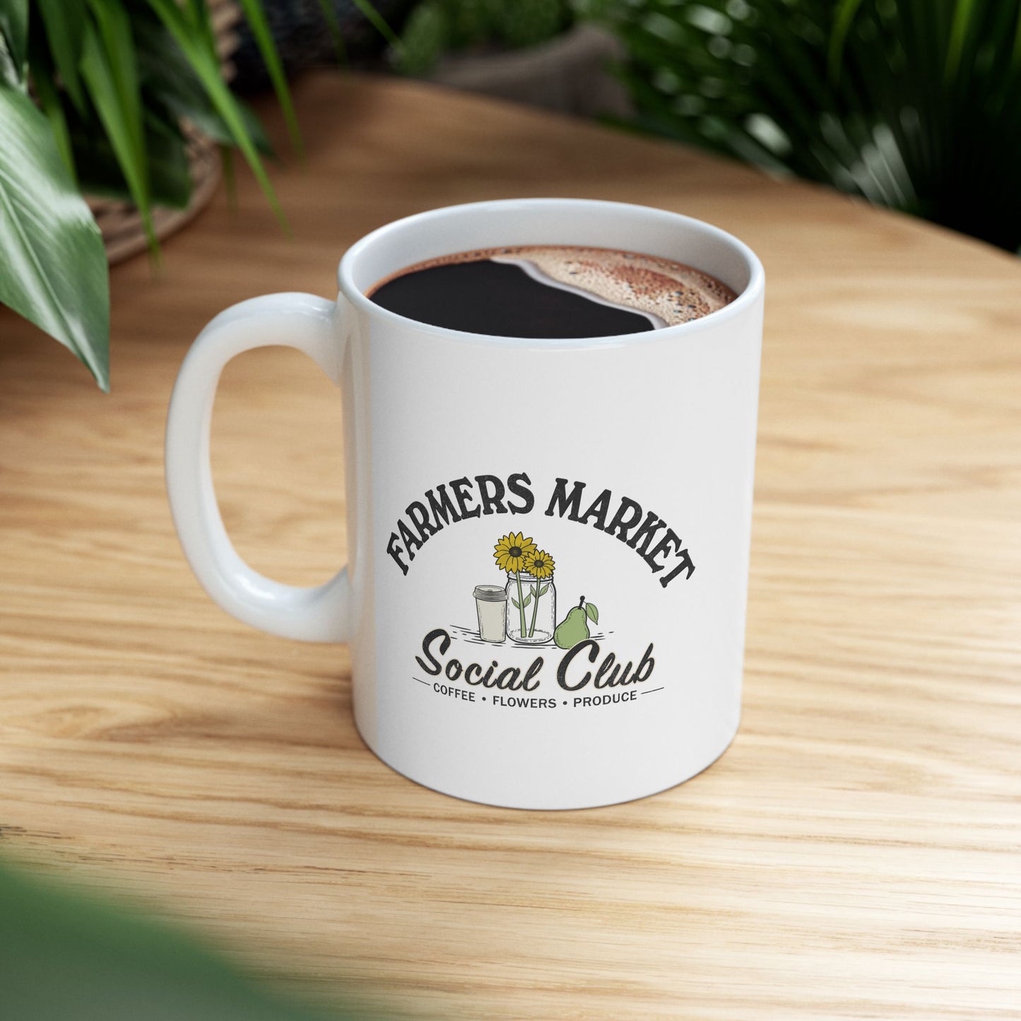 A white ceramic coffee mug with a black and green graphic reading 'FARMERS MARKET Social Club' sits on a light wood table with blurred green plants in the background.