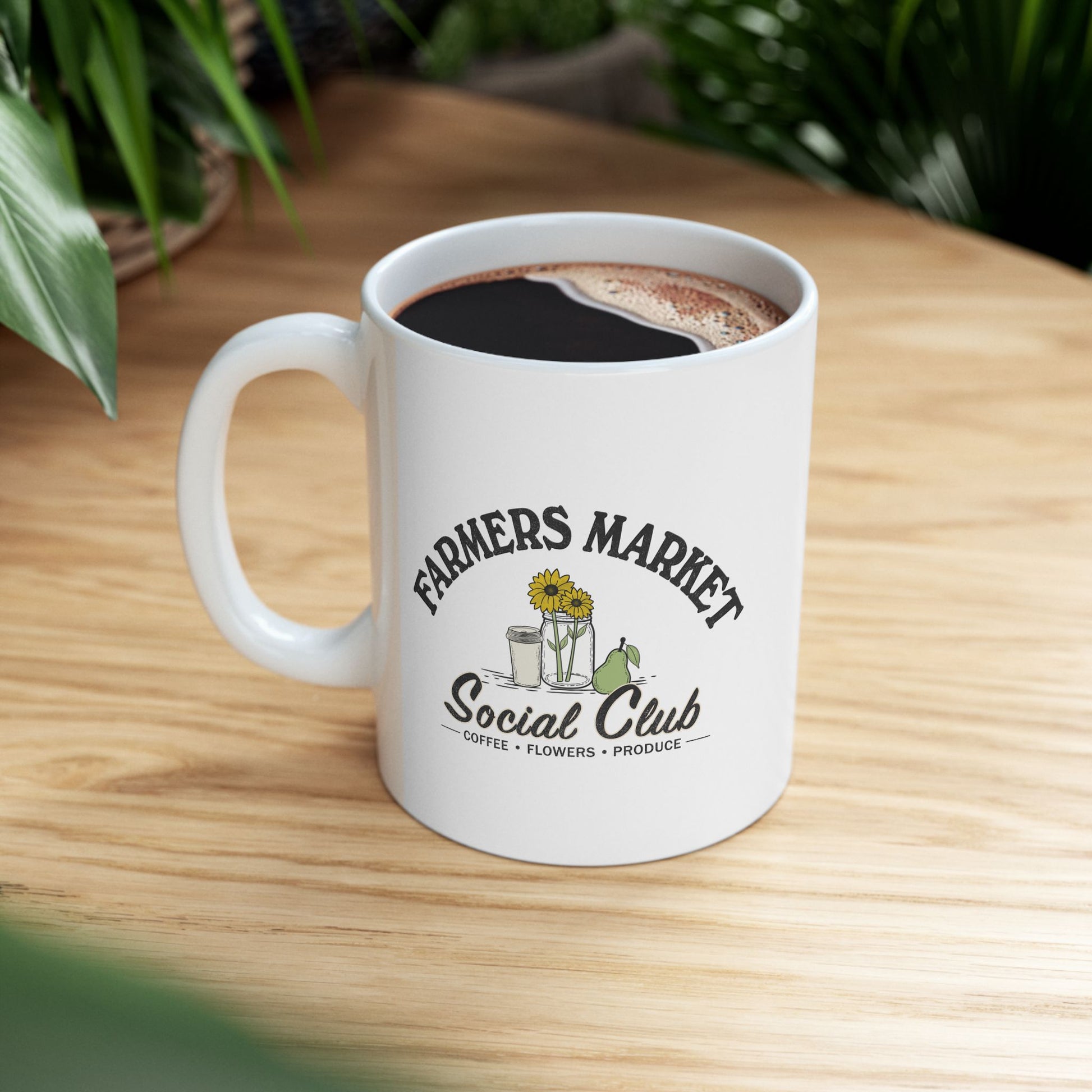 A white ceramic coffee mug with a black and green graphic reading 'FARMERS MARKET Social Club' sits on a light wood table with blurred green plants in the background.