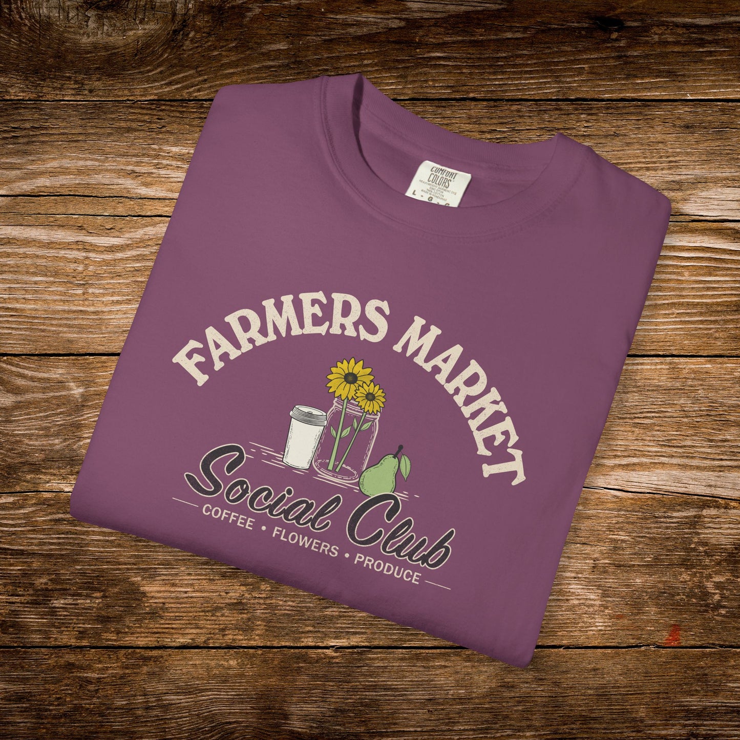 A folded purple t-shirt with a 'Farmers Market Social Club' graphic, displayed on a rustic wooden background.