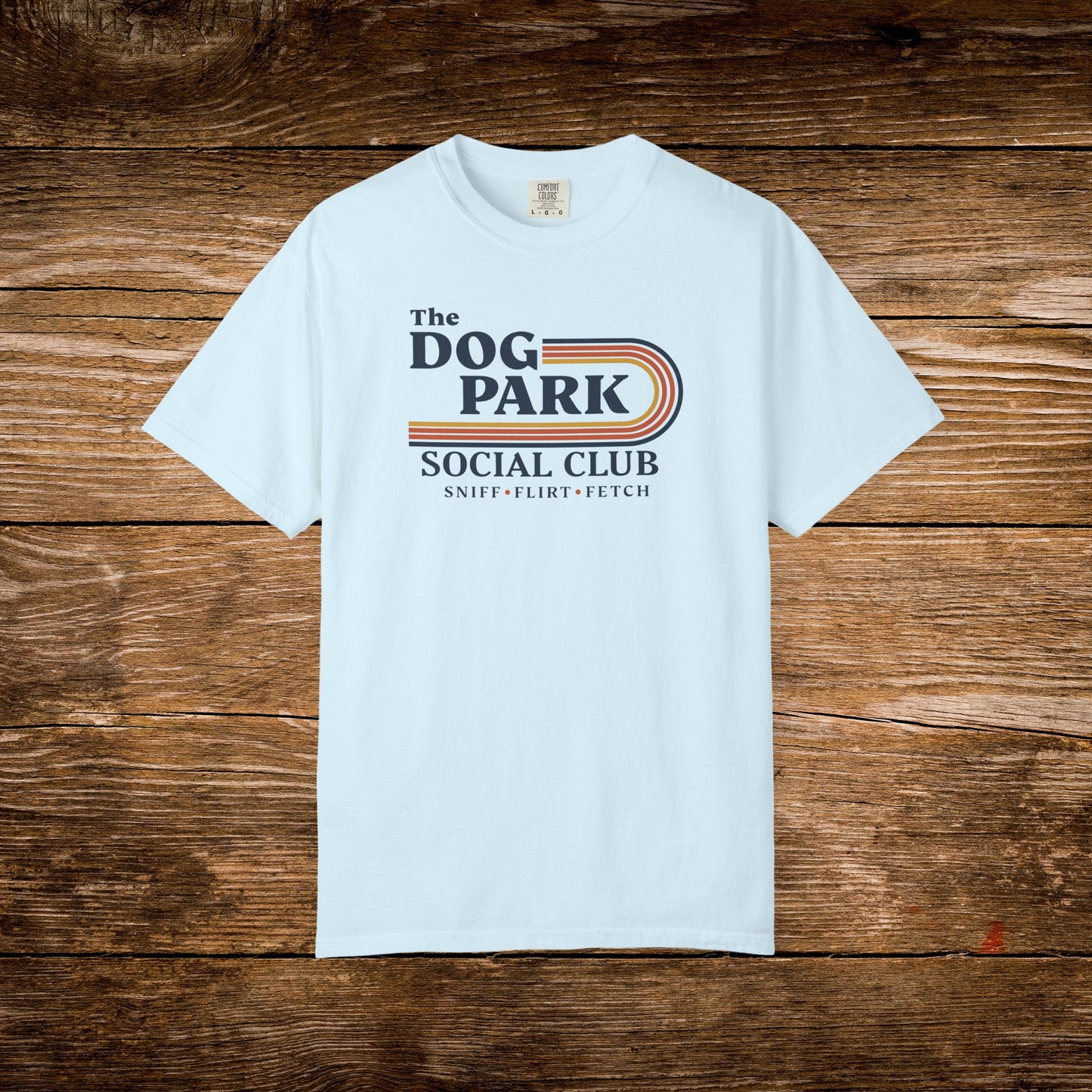 A light blue crewneck t-shirt with a retro-style graphic reading 'The DOG PARK SOCIAL CLUB' and 'SNIFF - FLIRT - FETCH' in navy, orange, and beige, laid flat on a rustic brown wooden background. The design text reads "Sniff, Flirt, Fetch."