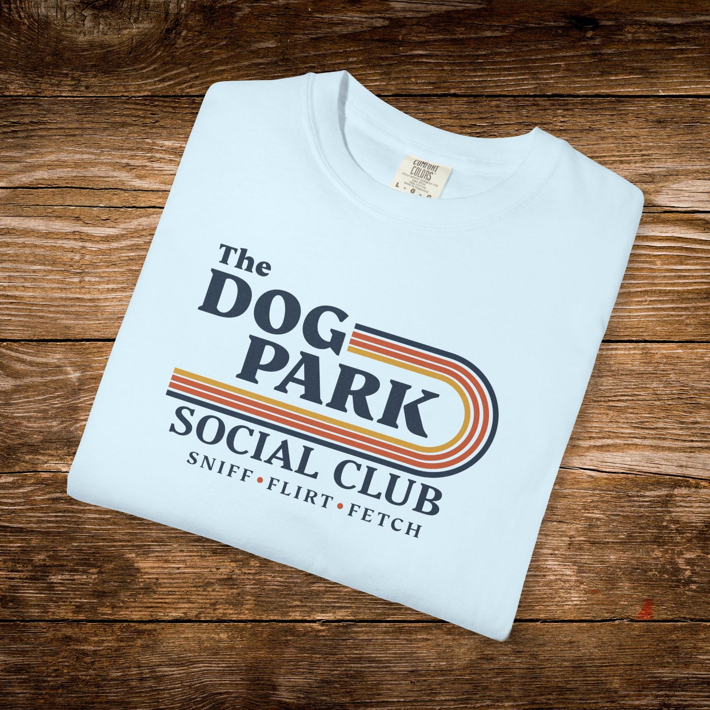A folded light blue t-shirt with a retro-style graphic reading 'The DOG PARK SOCIAL CLUB' and 'SNIFF • FLIRT • FETCH' in dark blue text with orange and yellow stripes, displayed on a rustic brown wooden background. The design text reads "Sniff, Flirt, Fetch."