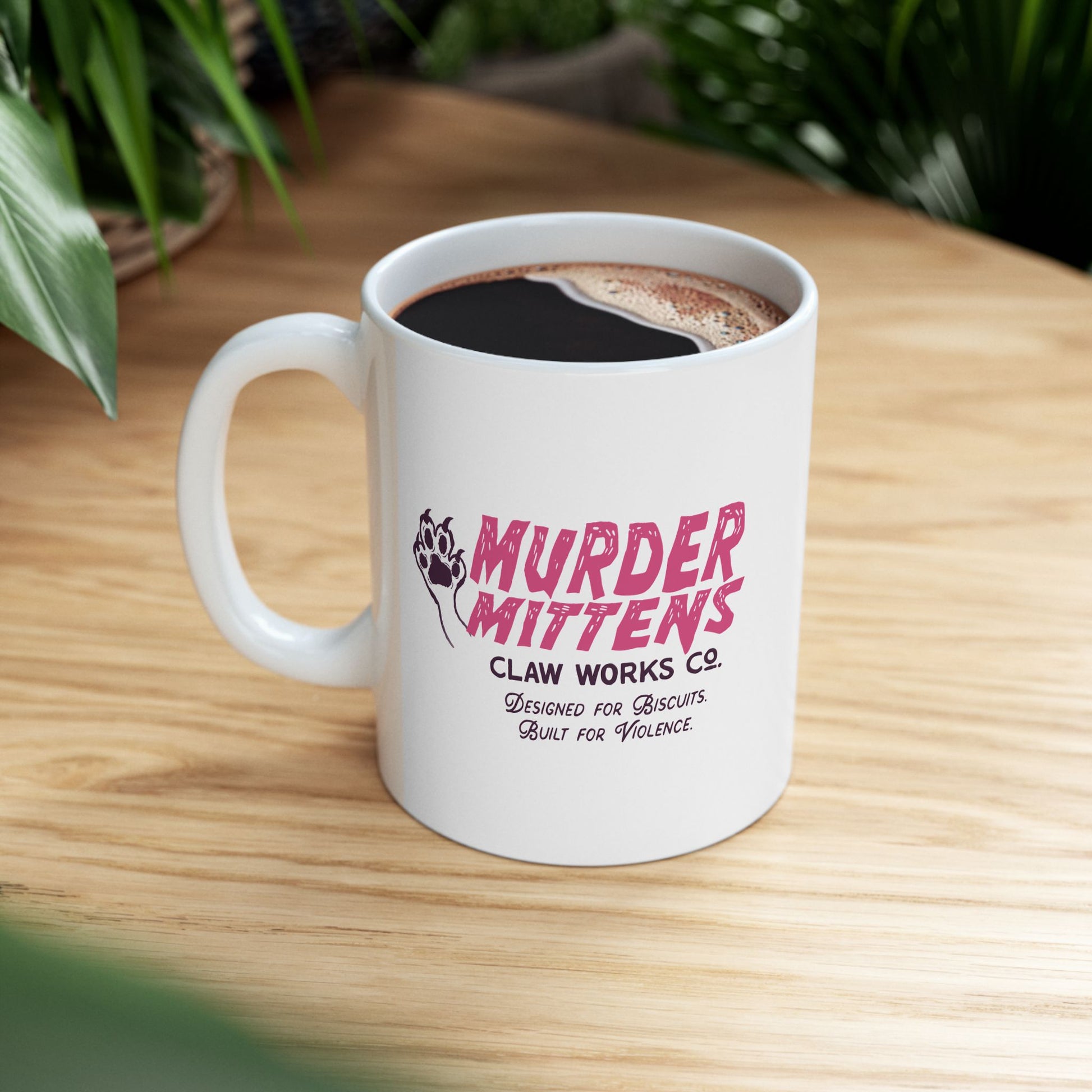 A white ceramic coffee mug with a pink and black 'Murder Mittens' graphic, filled with black coffee and sitting on a light wood tabletop with green plants in the background.