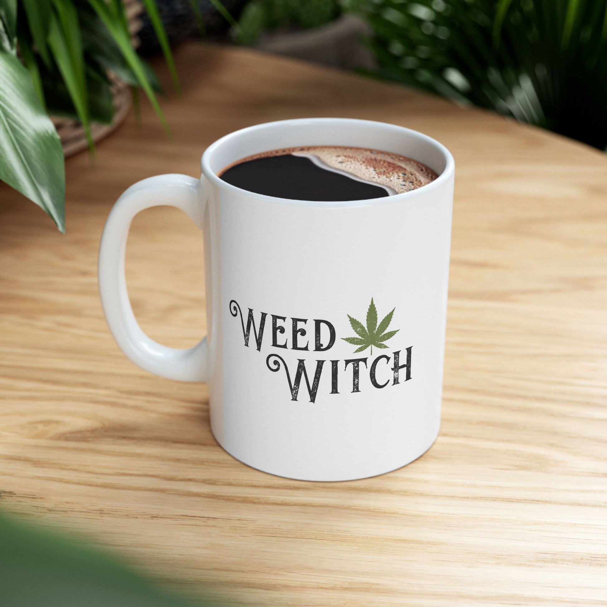 A white ceramic coffee mug with the words 'WEED WITCH' and a green cannabis leaf printed on the side, filled with black coffee and placed on a light wood table with blurred green plants in the background.
