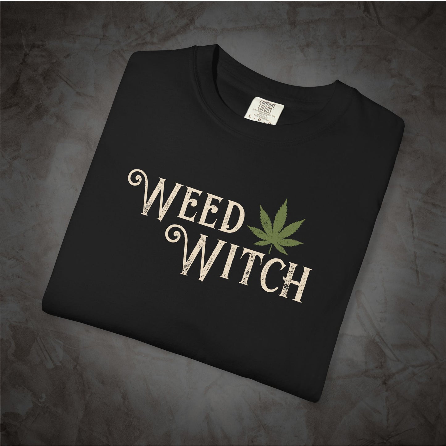 A folded black t-shirt with a distressed cream-colored 'WEED WITCH' graphic and a green cannabis leaf, displayed on a dark textured background.