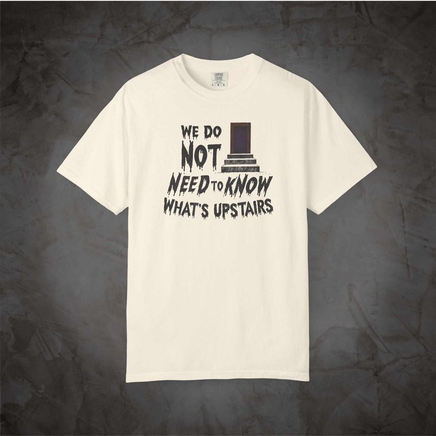 An off-white short-sleeve t-shirt with a black graphic print on the front reading 'WE DO NOT NEED TO KNOW WHAT'S UPSTAIRS' alongside a top hat illustration, displayed against a dark textured background.