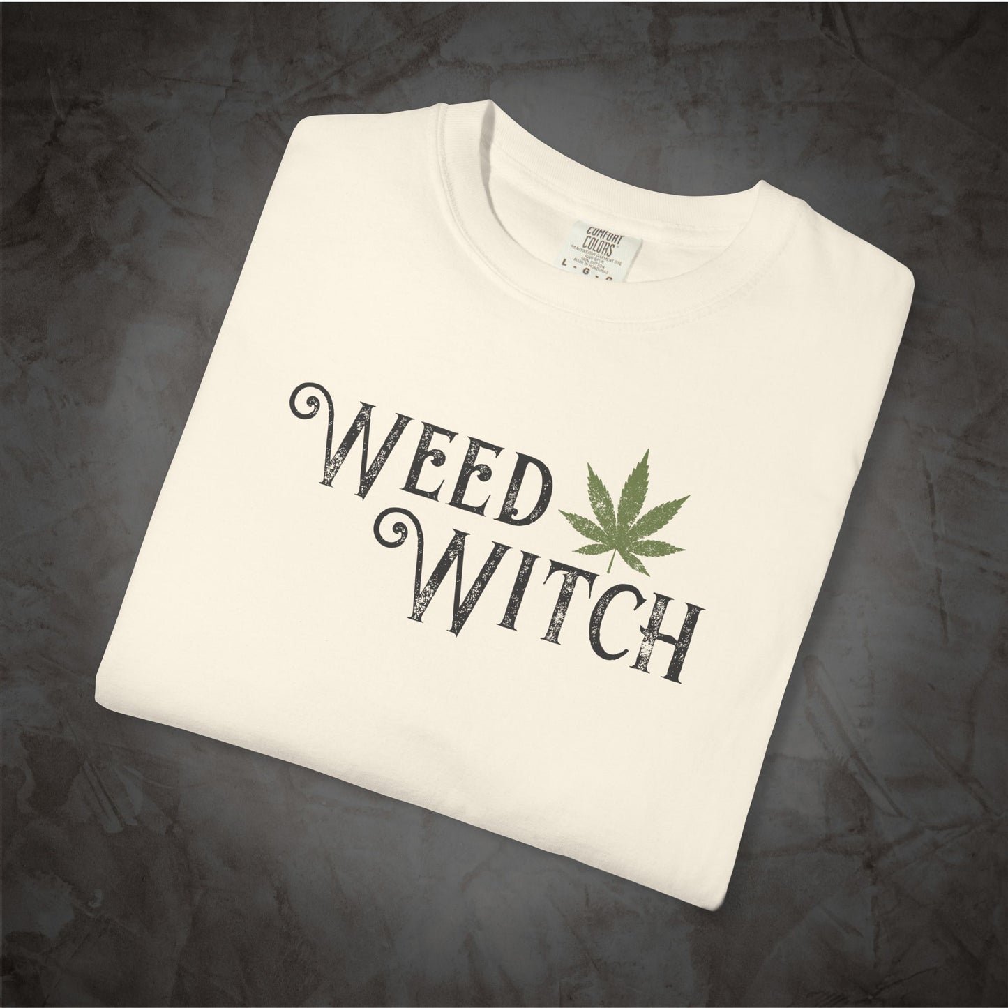 A folded off-white t-shirt with a graphic print reading 'WEED WITCH' in a distressed black font, accented by a green cannabis leaf, displayed on a dark textured background.