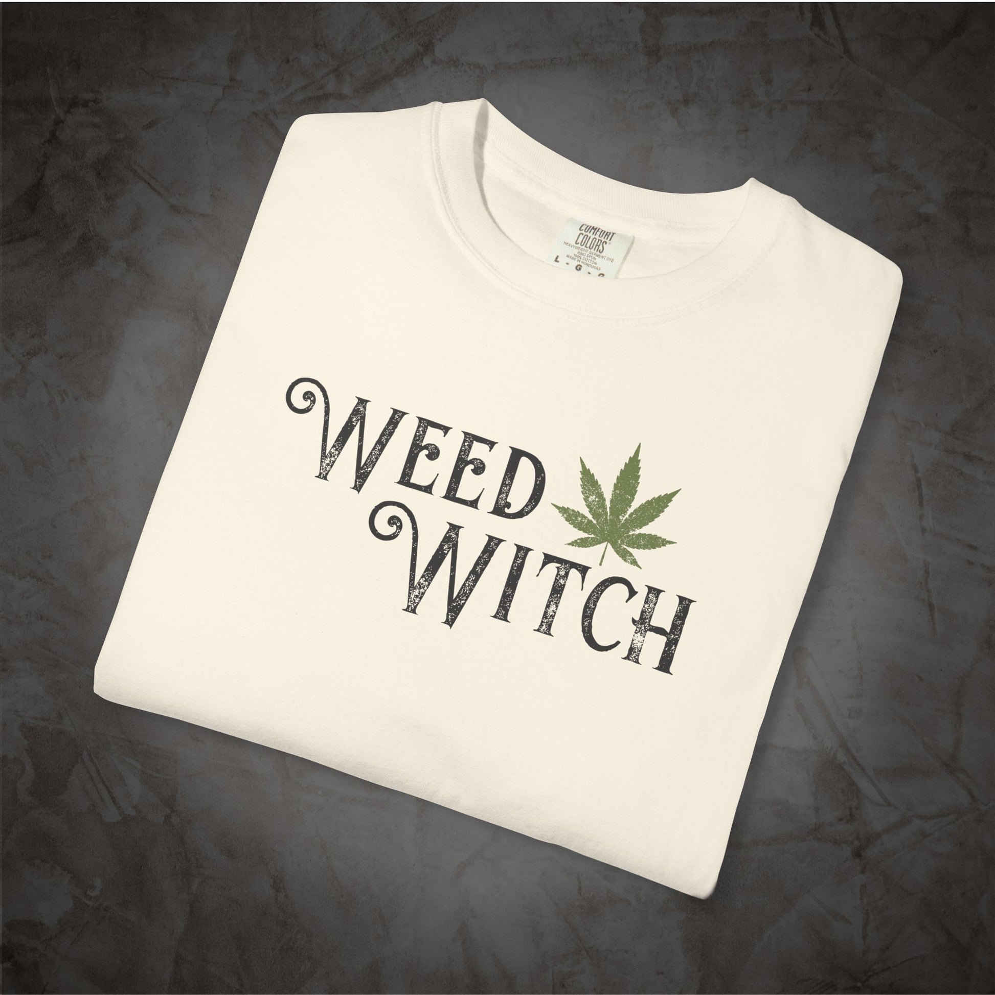 A folded off-white t-shirt with a graphic print reading 'WEED WITCH' in a distressed black font, accented by a green cannabis leaf, displayed on a dark textured background.