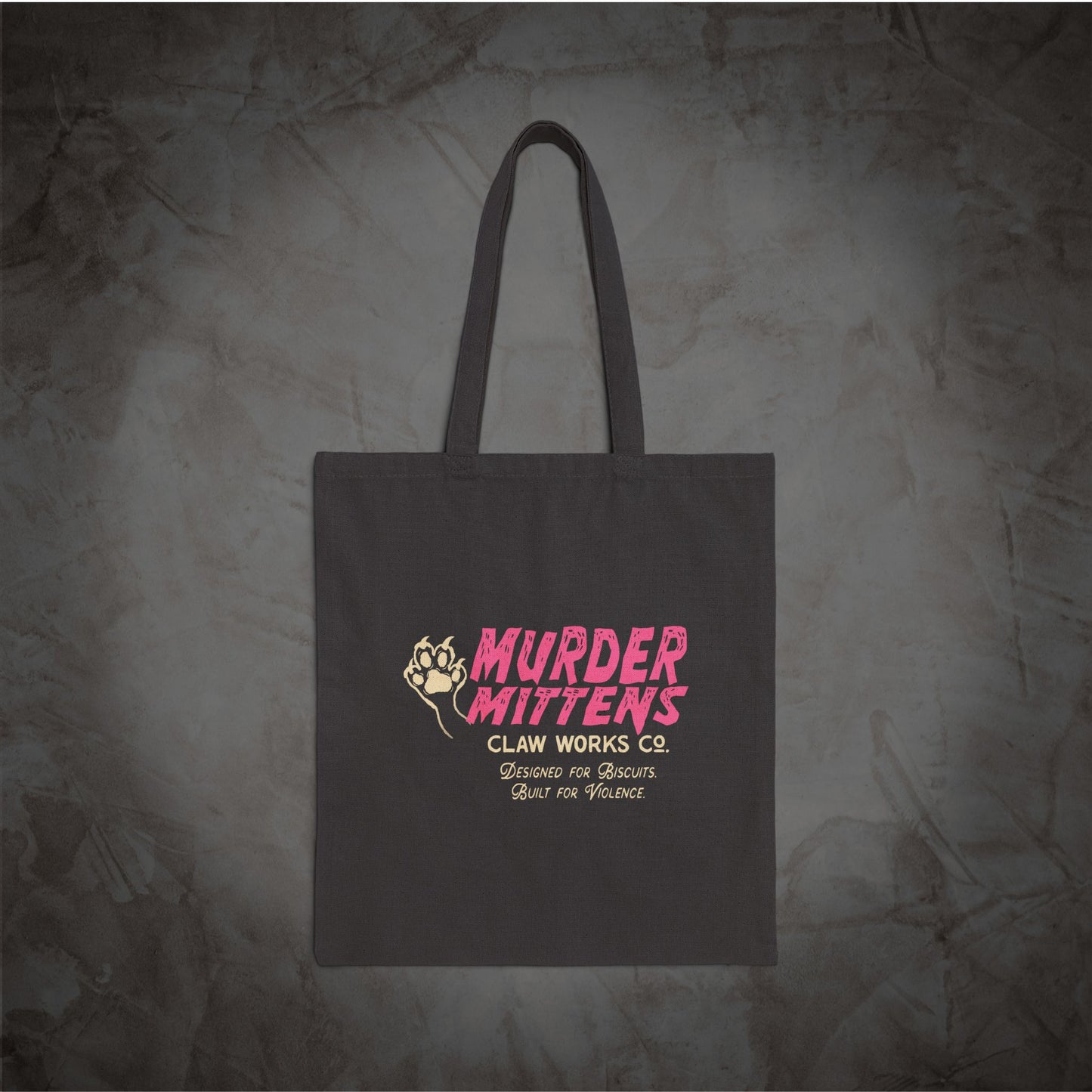 A dark gray canvas tote bag with long handles, featuring a pink and yellow graphic print that reads 'MURDER MITTENS CLAW WORKS Co.' with smaller text below, displayed against a textured gray background.