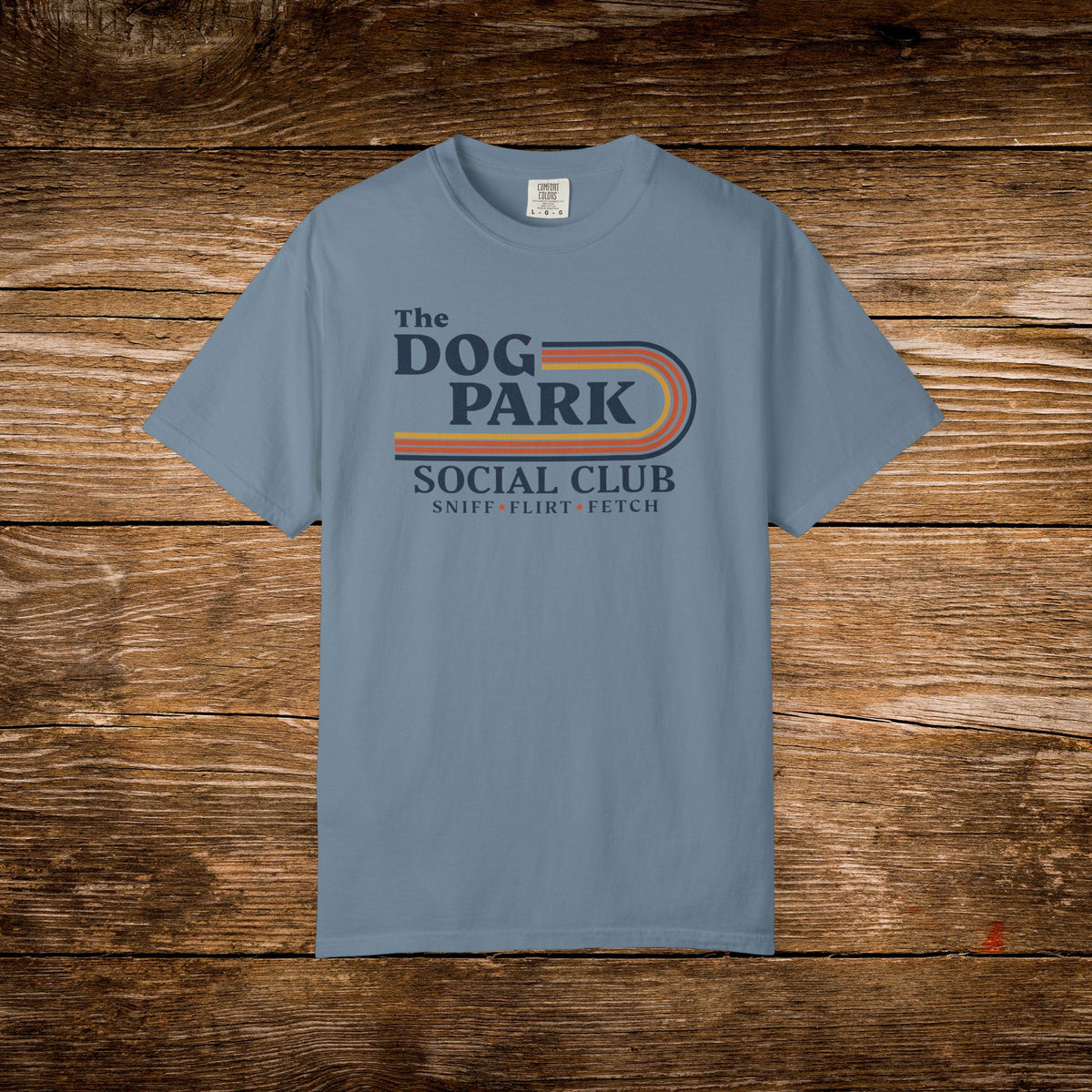 A light blue crewneck t-shirt with a retro-style graphic reading 'The DOG PARK SOCIAL CLUB' and 'SNIP-F FLIRT-FETCH' underneath, displayed flat against a rustic brown wooden plank background.