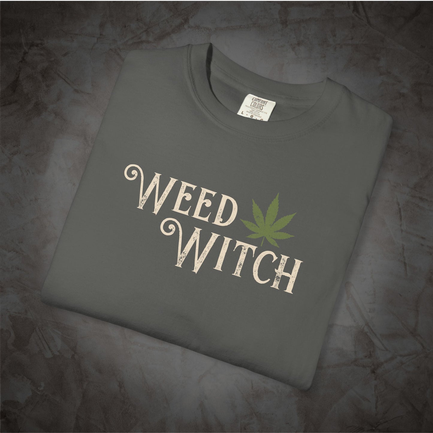 A folded dark gray t-shirt with a distressed cream-colored 'WEED WITCH' graphic and a green cannabis leaf, displayed on a textured gray background.