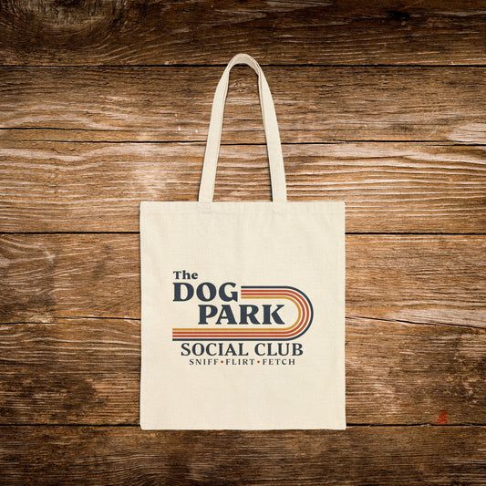 A cream canvas tote bag with 'The Dog Park Social Club' printed on the front, displayed flat against a rustic brown wooden plank background.