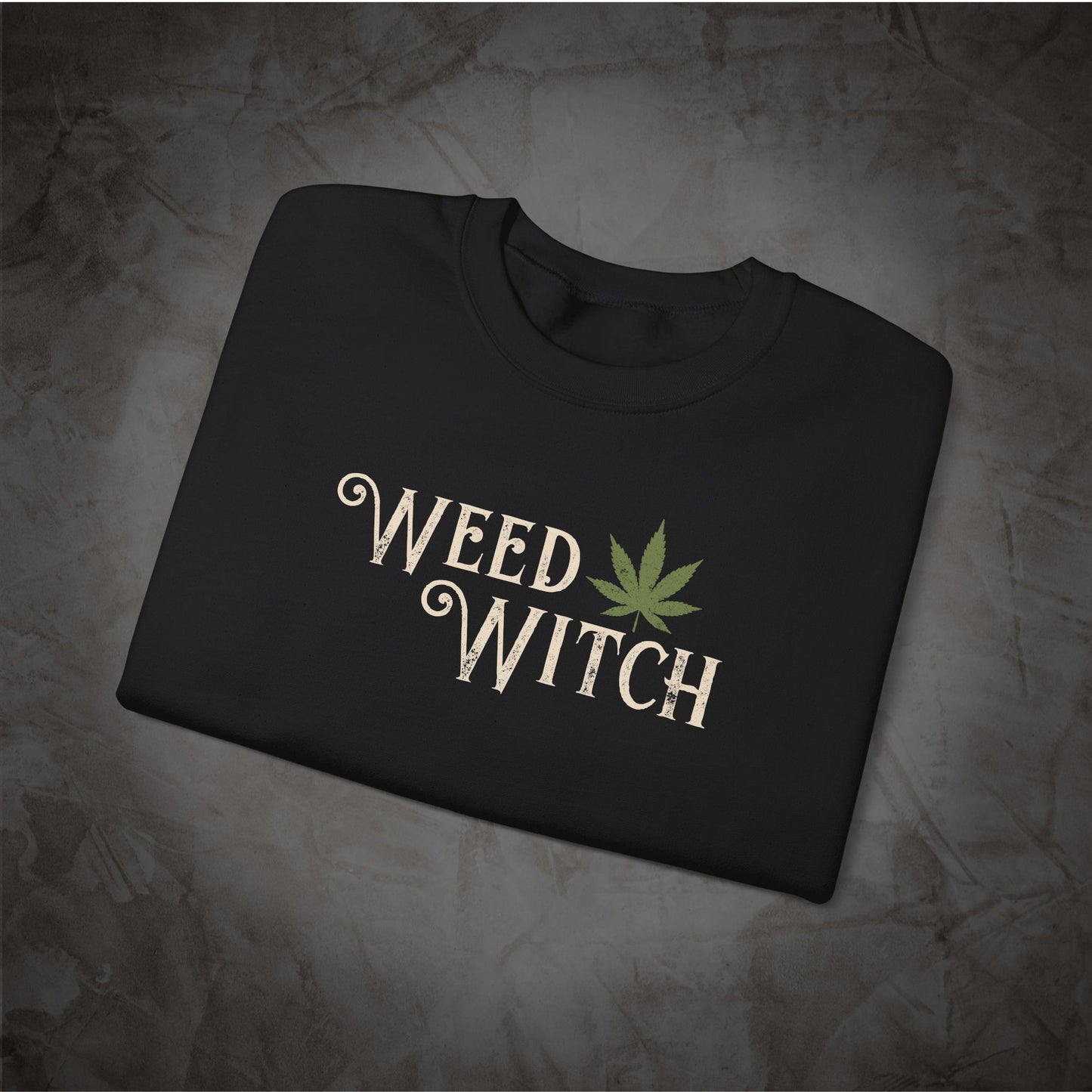 A folded black crewneck t-shirt with a graphic print reading 'WEED WITCH' in a beige gothic font next to a green cannabis leaf, displayed on a textured gray background.