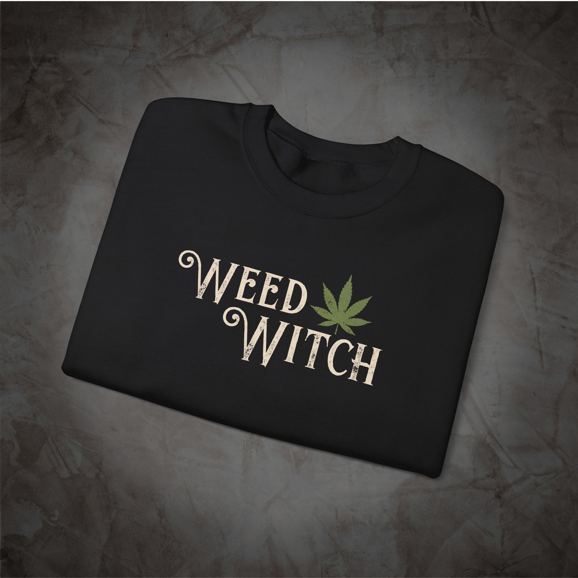 A folded black crewneck t-shirt with a graphic print reading 'WEED WITCH' in a beige gothic font next to a green cannabis leaf, displayed on a textured gray background.