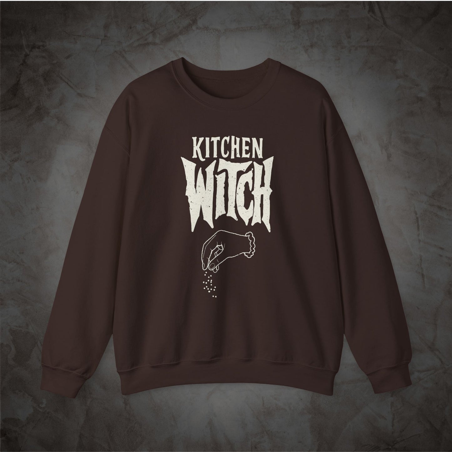 A dark brown crewneck sweatshirt featuring a white graphic print that reads 'KITCHEN WITCH' above an illustration of a hand sprinkling spices, displayed against a textured gray background.