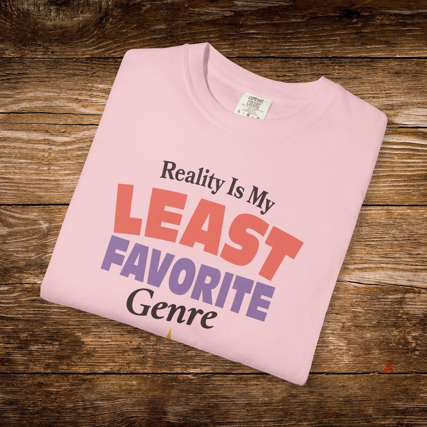 A folded light pink t-shirt with the text 'Reality Is My LEAST FAVORITE Genre' printed on the front, displayed on a rustic wooden background.
