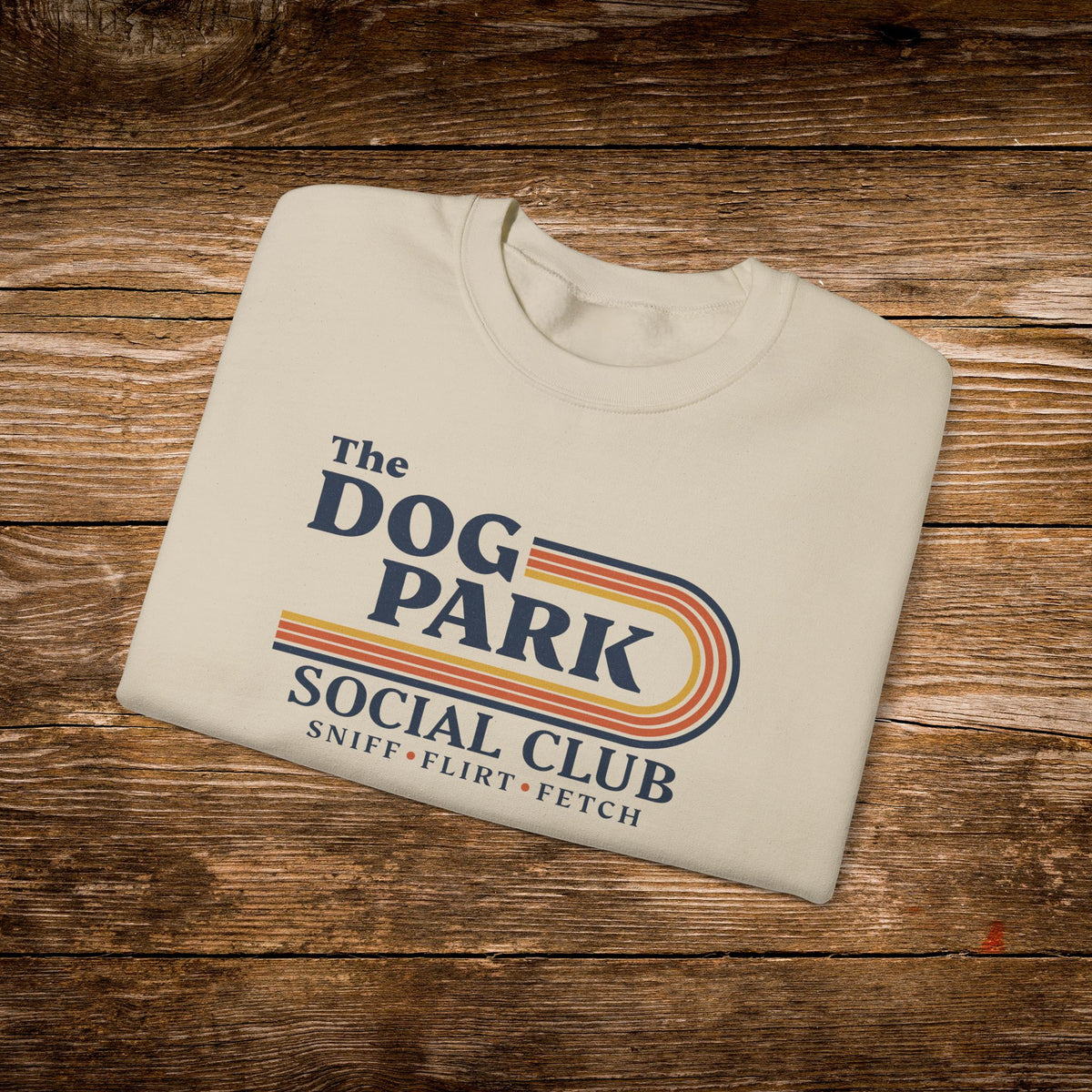 A folded cream-colored t-shirt with a retro-style graphic reading 'The DOG PARK SOCIAL CLUB' and 'SNIFF • FLIRT • FETCH' in navy, orange, and yellow, displayed on a rustic wooden background.