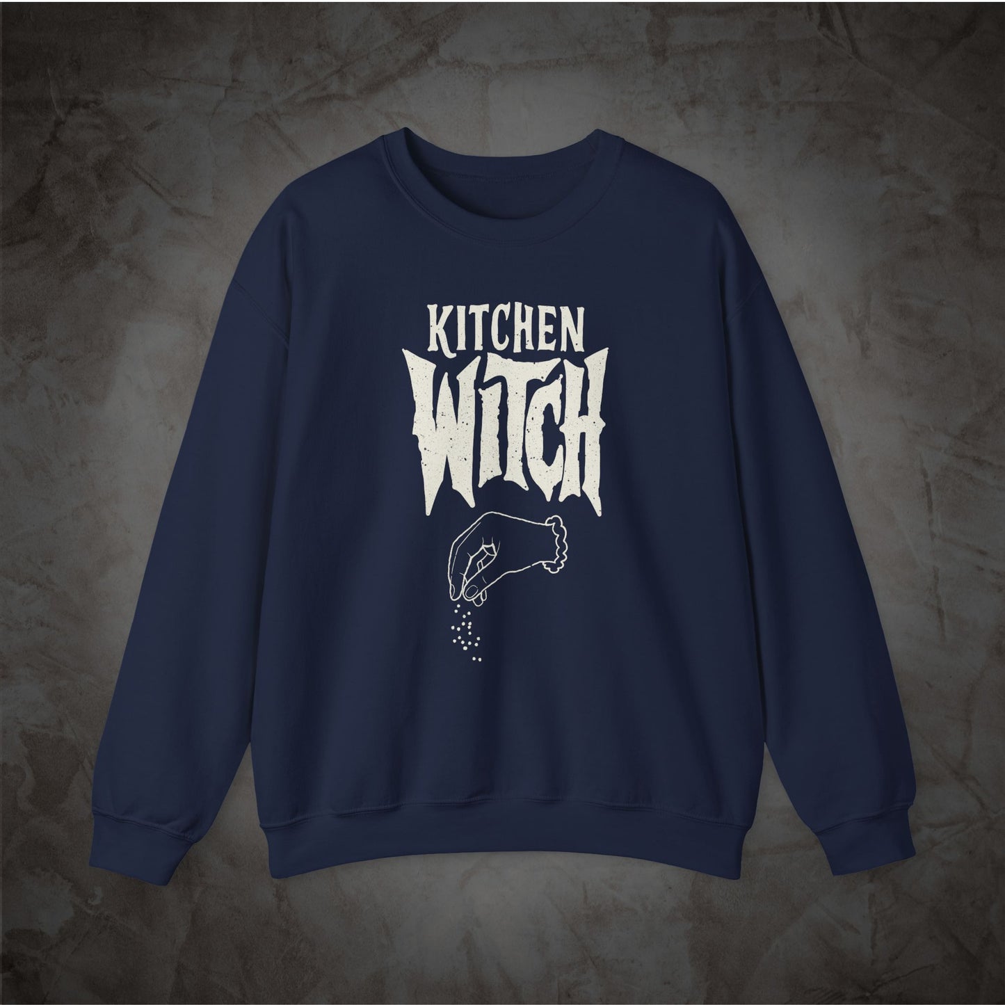 A navy blue crewneck sweatshirt with a white graphic print reading 'KITCHEN WITCH' and a hand sprinkling seasoning, displayed against a textured gray background.