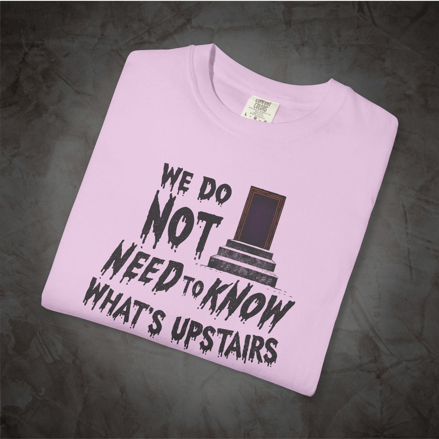 A light pink folded t-shirt with a black graphic print that reads 'WE DO NOT NEED TO KNOW WHAT'S UPSTAIRS' alongside a small illustration of a door, displayed on a dark textured background.