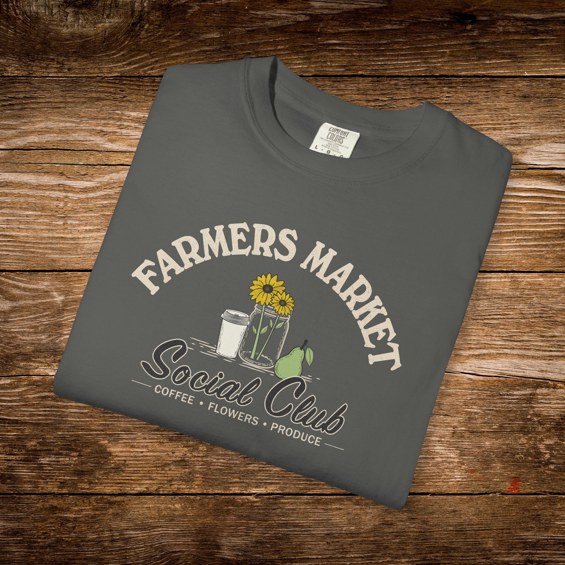 A dark gray folded t-shirt with a 'Farmers Market Social Club' graphic, displayed on a rustic wooden background.