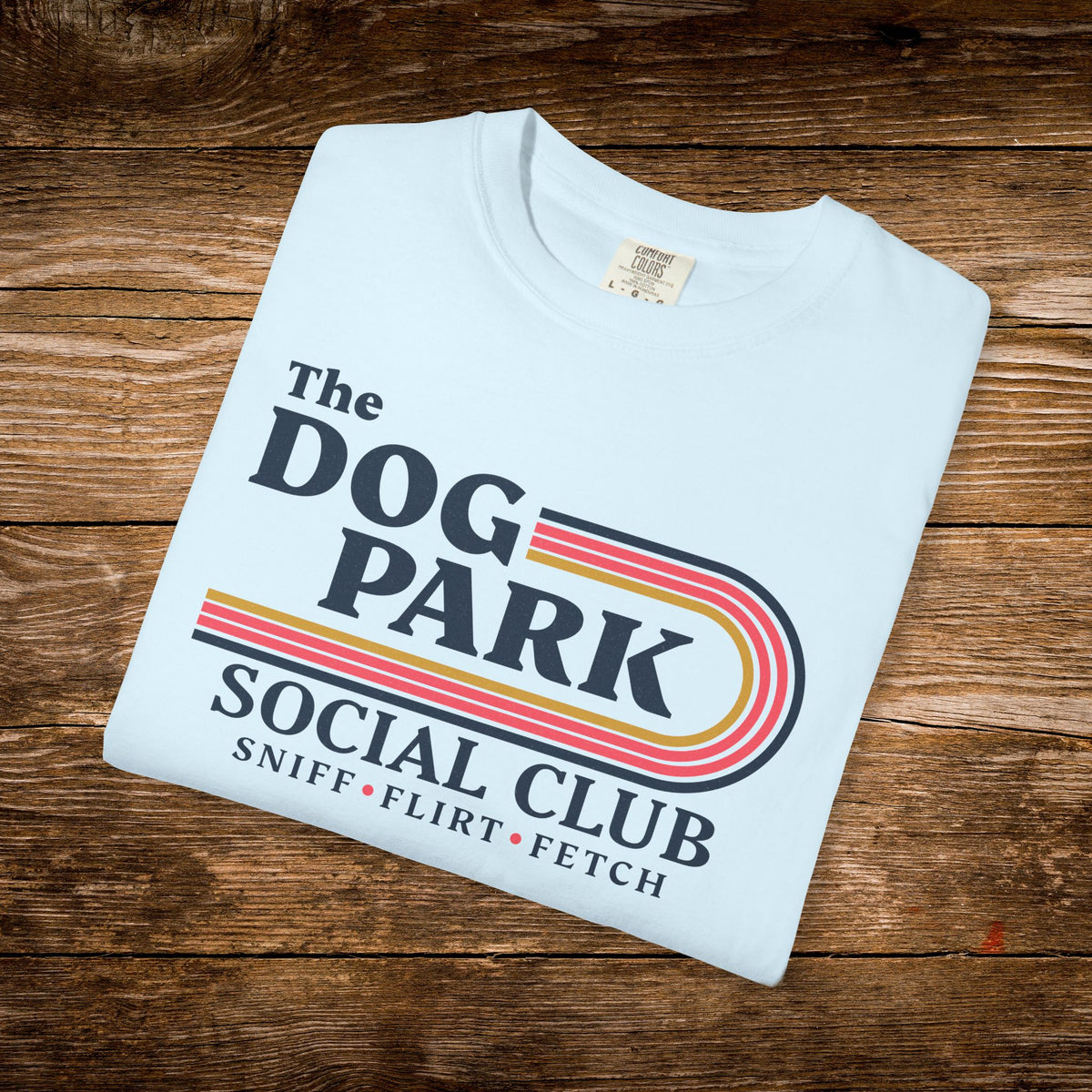 A folded off-white t-shirt with a retro-style 'The Dog Park Social Club' graphic, displayed on a rustic wooden plank background.