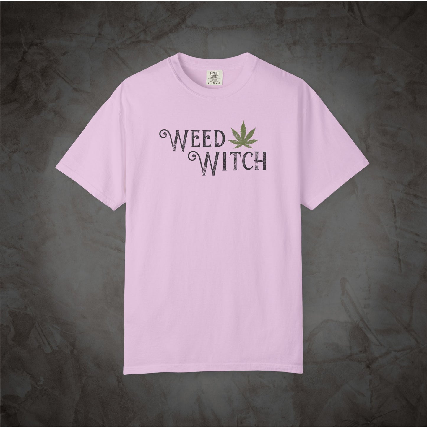 A light pink short-sleeve t-shirt with 'WEED WITCH' printed in a dark gray gothic font and a green cannabis leaf graphic, displayed flat against a dark textured gray background.