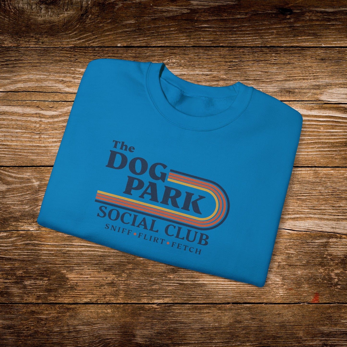 A folded bright blue t-shirt with a retro-style graphic reading 'The Dog Park Social Club' and 'Sniff-Flirt-Fetch' in dark blue text with a rainbow stripe, displayed on a rustic brown wooden background. The design text reads "Sniff, Flirt, Fetch."